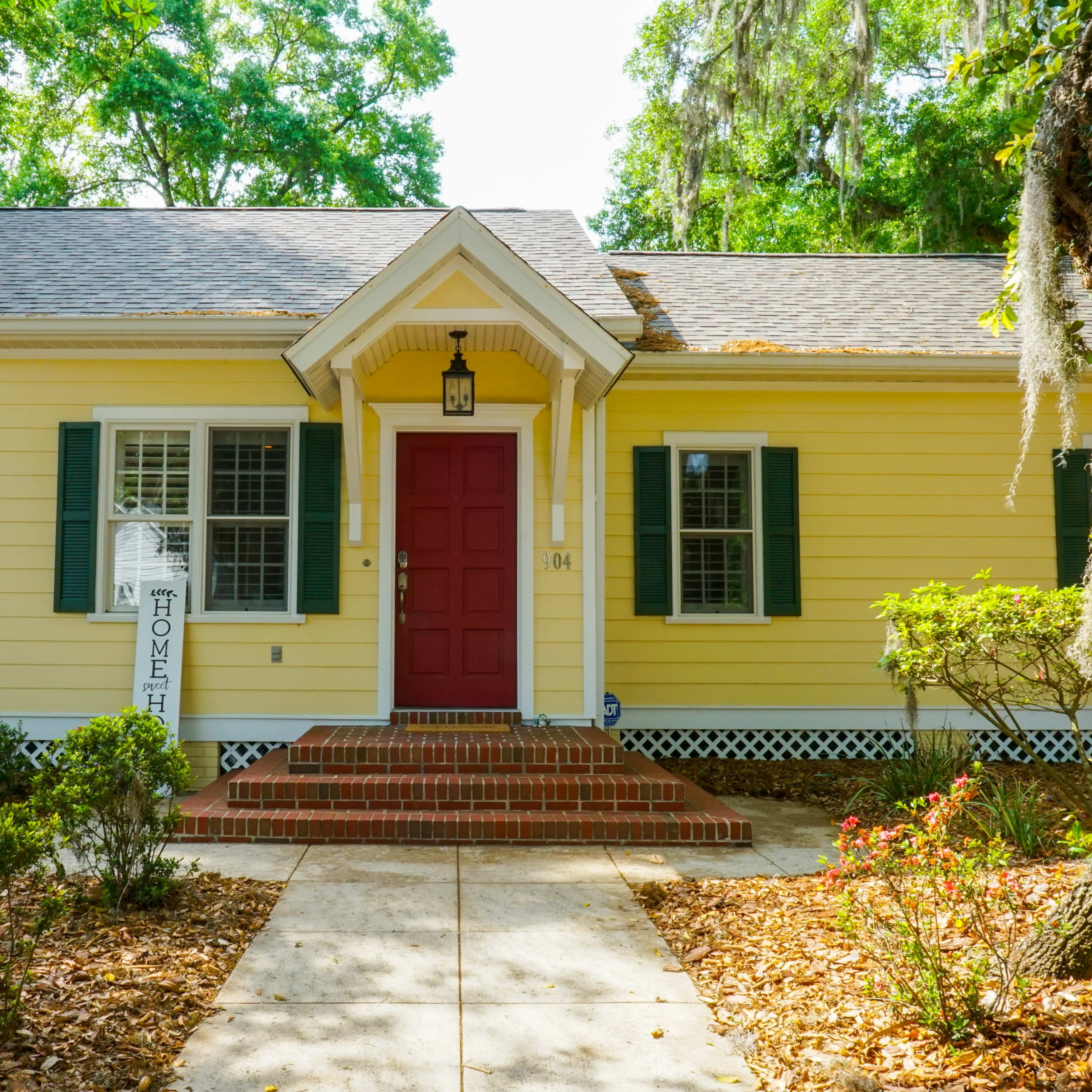 A yellow house with a red front door, white shutters, and a brick walkway leading to the entrance.