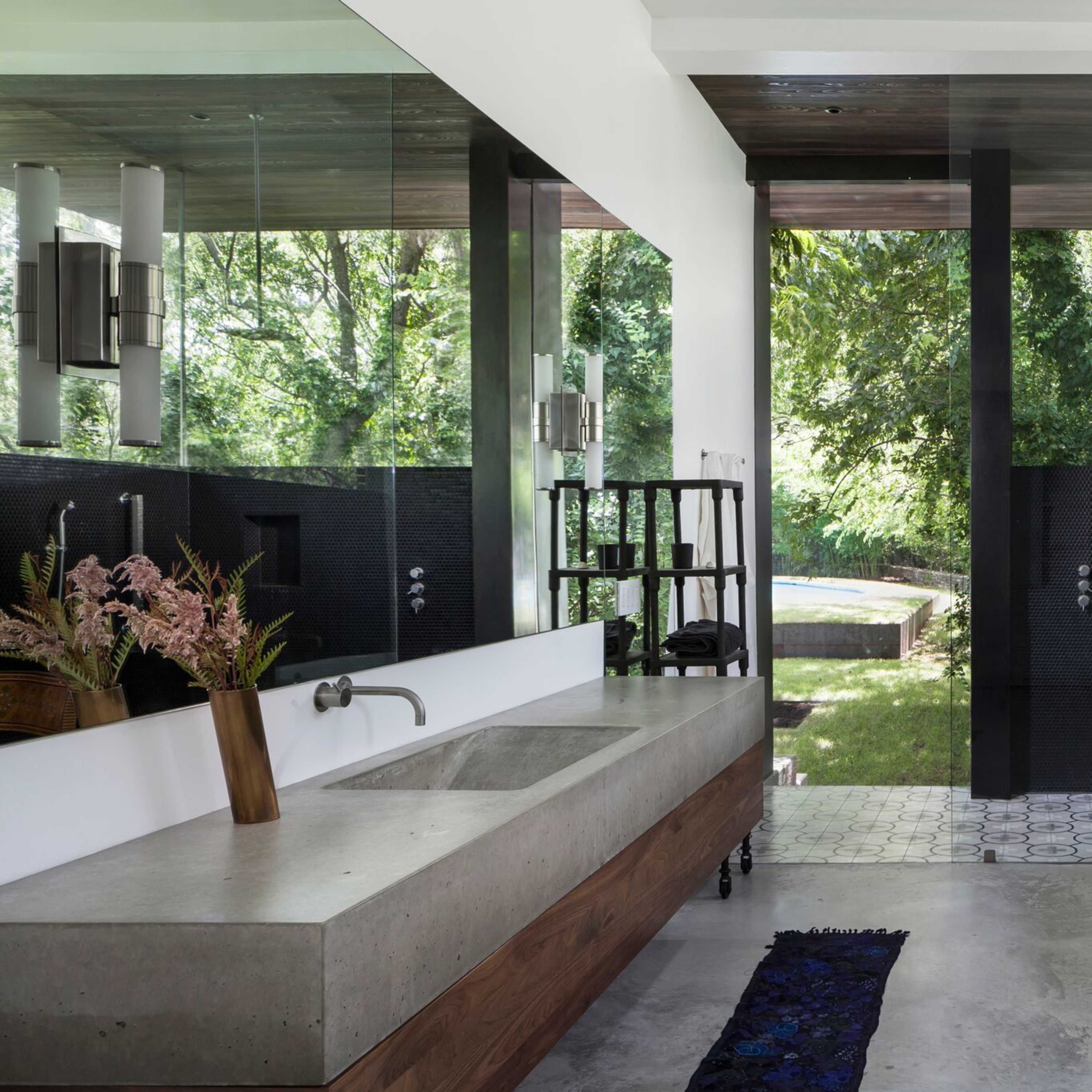 The bathroom features a concrete sink and tub, large mirrors, and an outdoor view framed by glass walls.