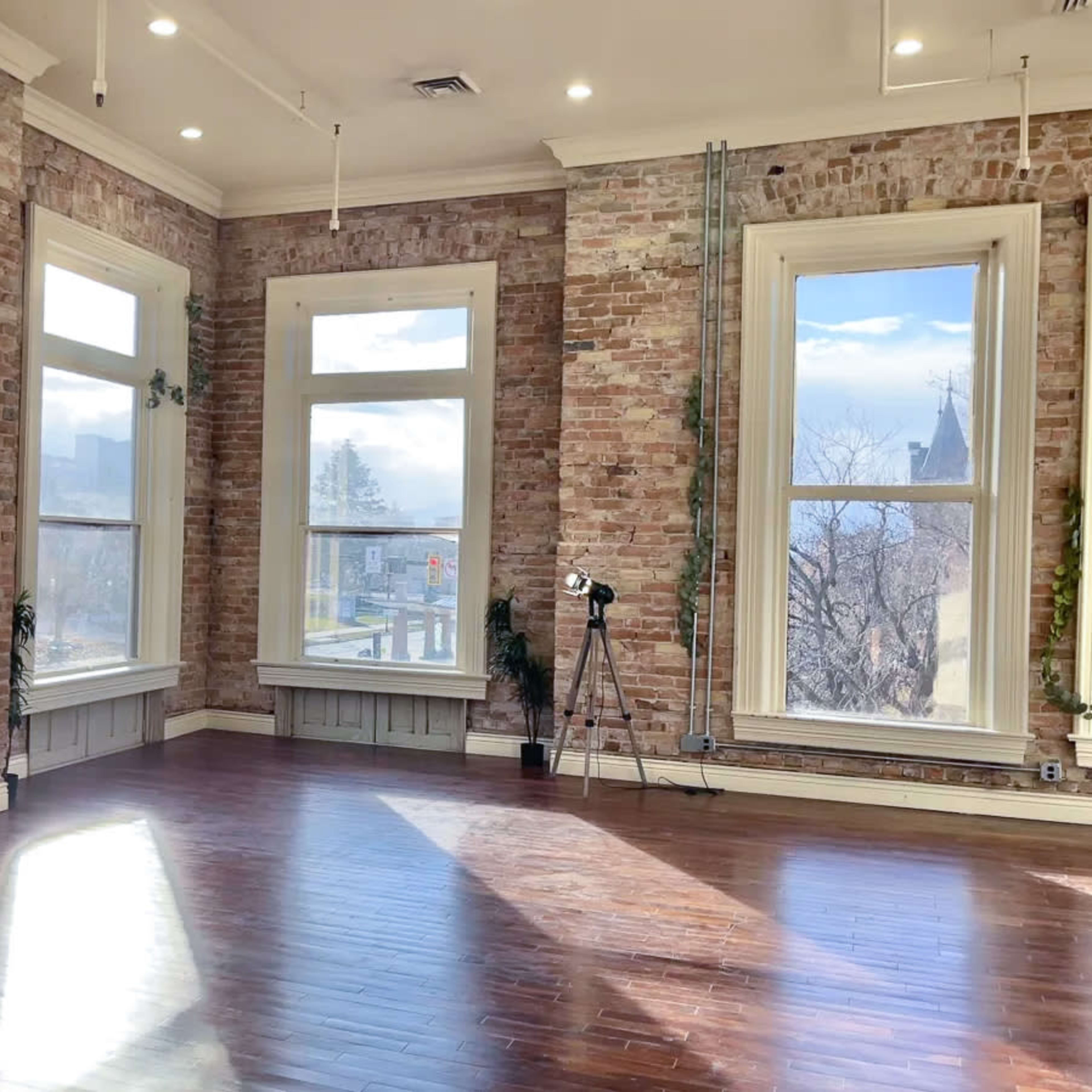 An empty room with exposed brick walls, large windows, and wooden flooring.
