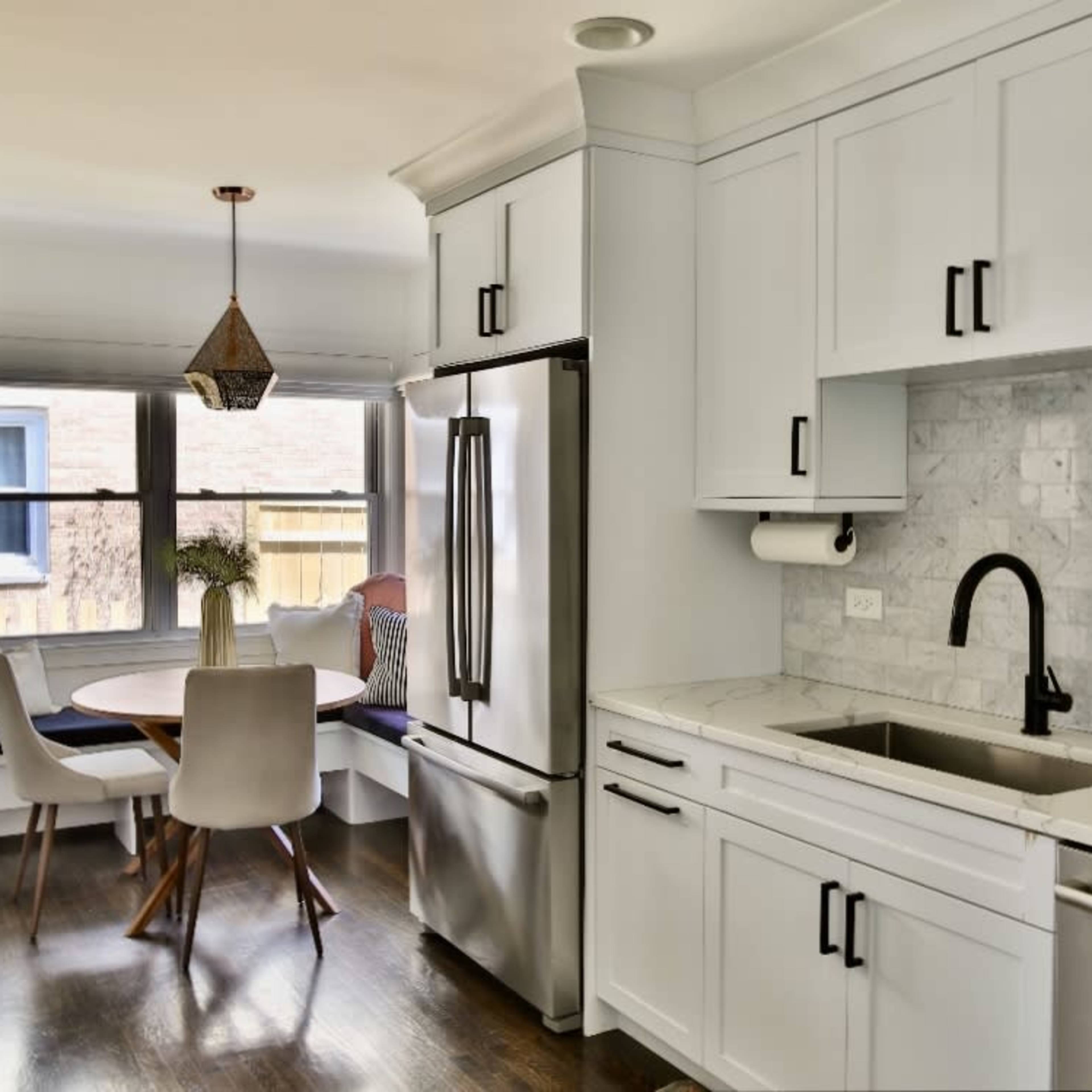 A modern kitchen features a stainless steel refrigerator, a marble countertop, a sink, and a circular dining table surrounded by chairs near a window bench.