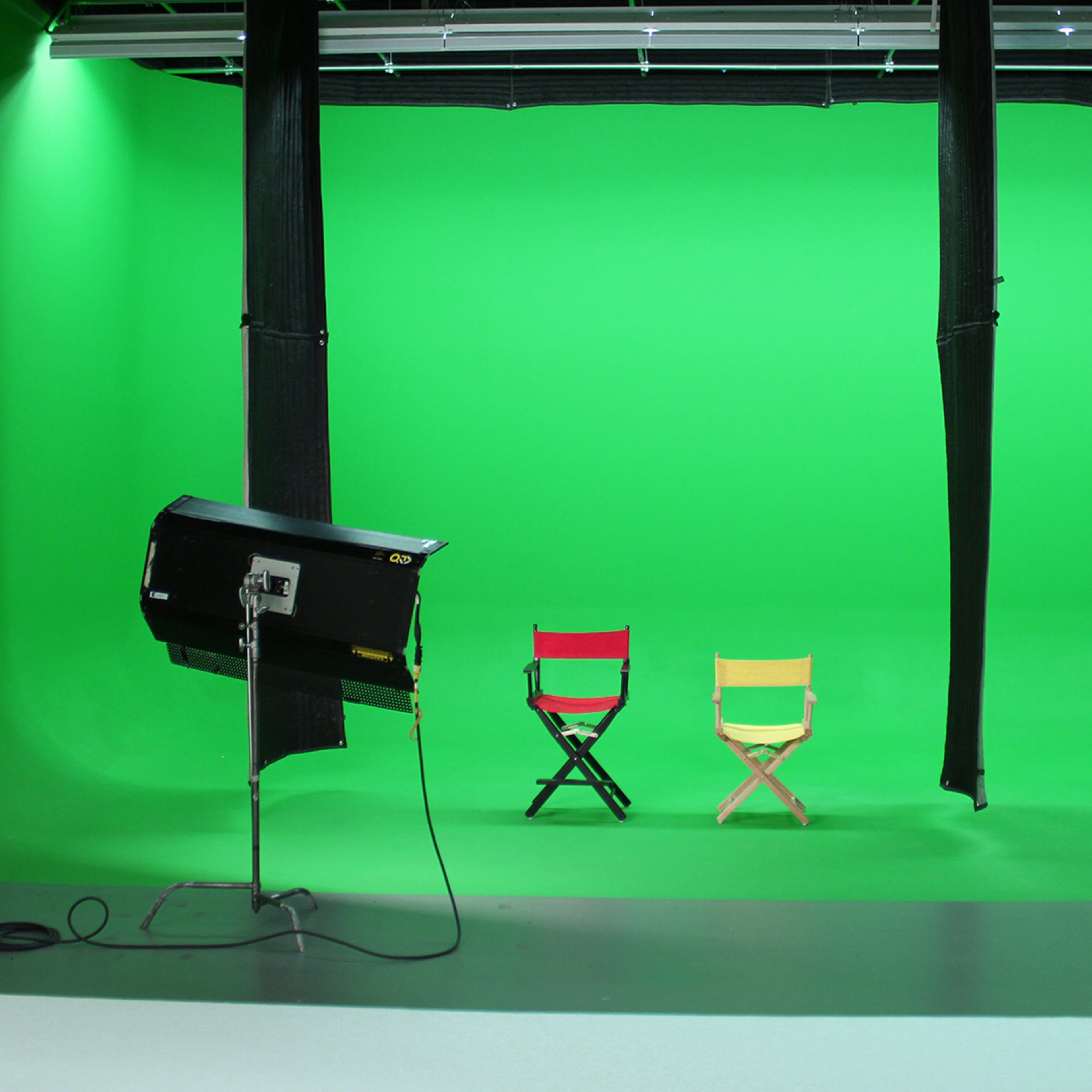 A photography studio with a green screen backdrop and two empty directors' chairs, one red and one yellow, positioned in front.