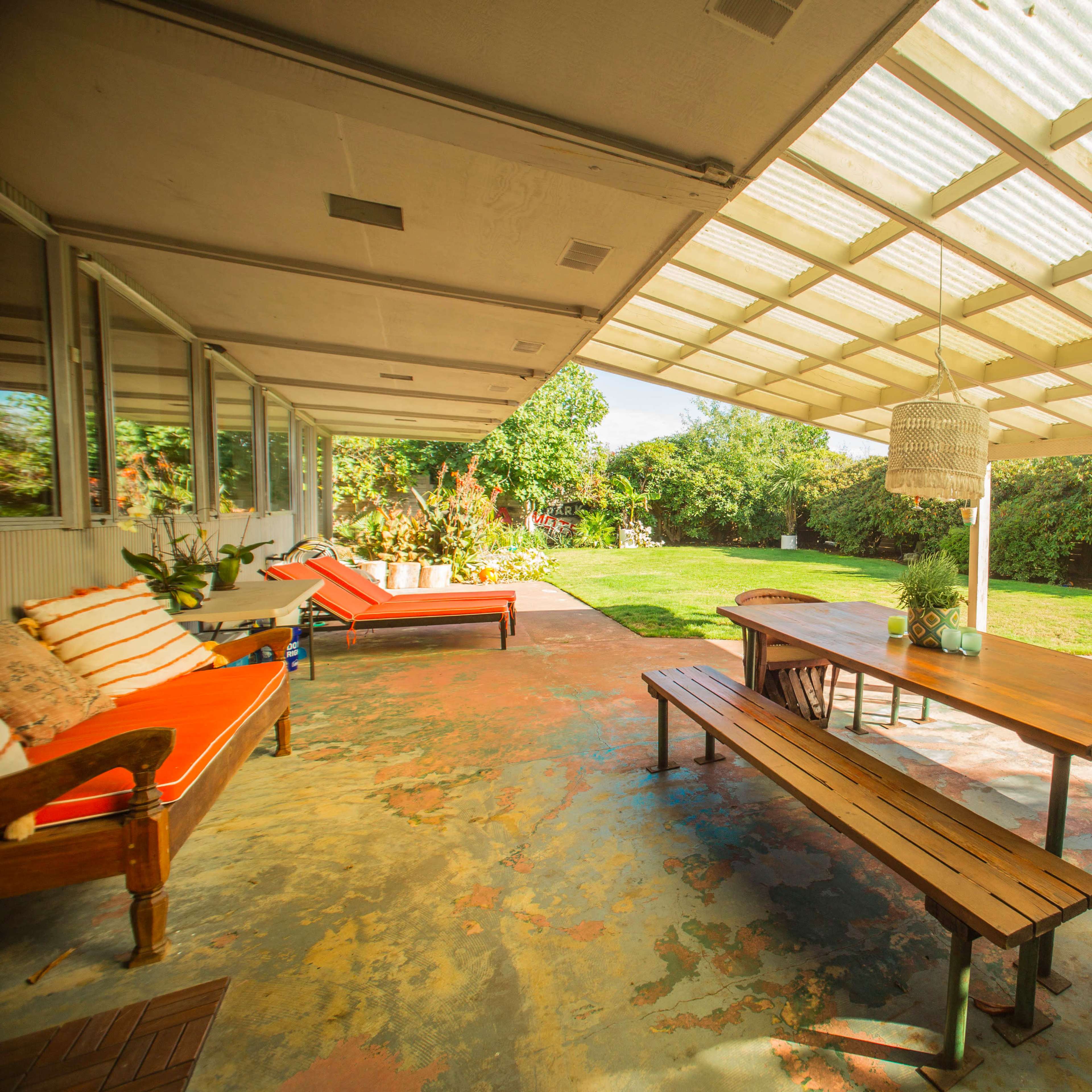 A covered patio features a wooden table and benches, surrounded by a grassy yard and colorful flooring.