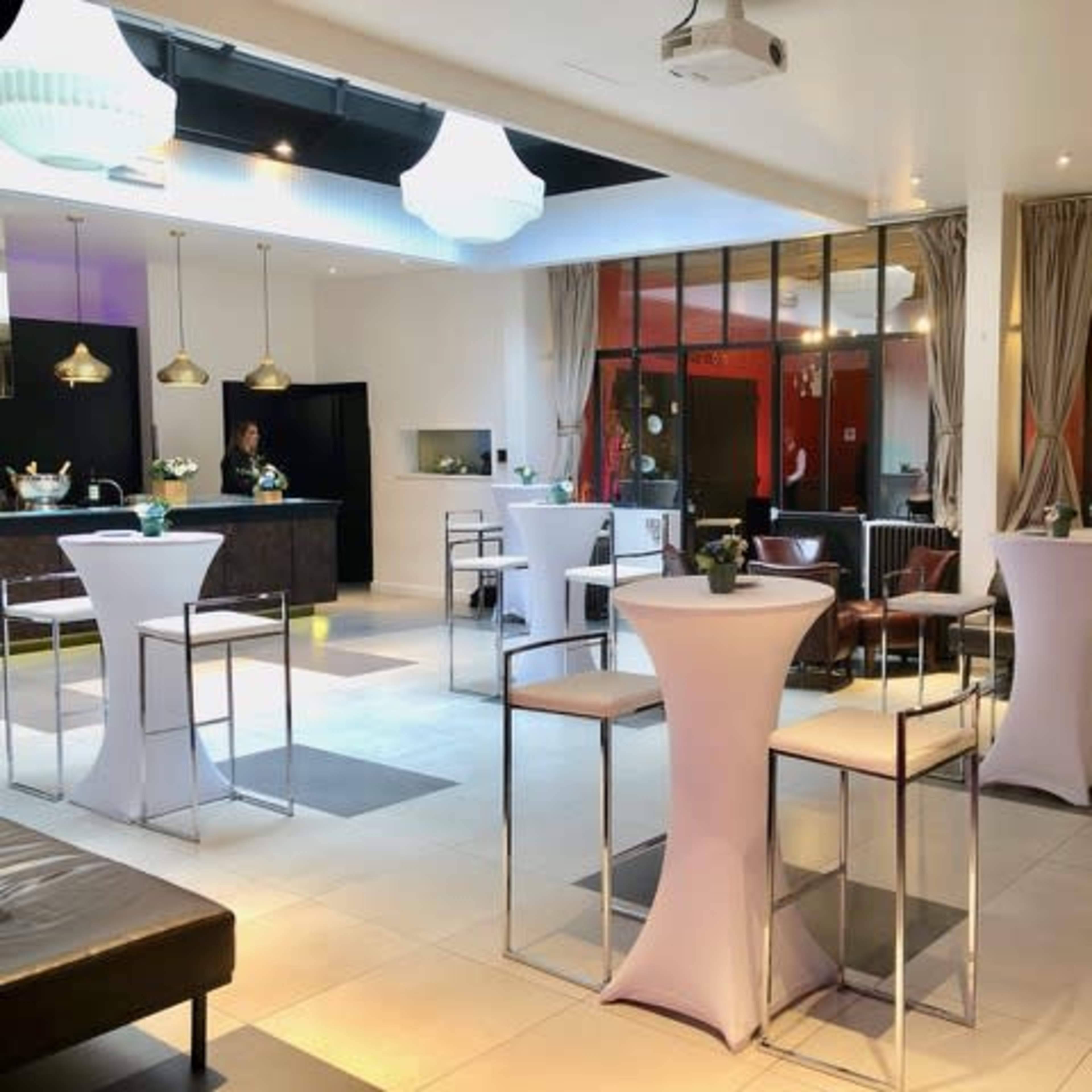 A modern event space with high tables and chairs, a bar area, and large windows providing a view of a lounge area.