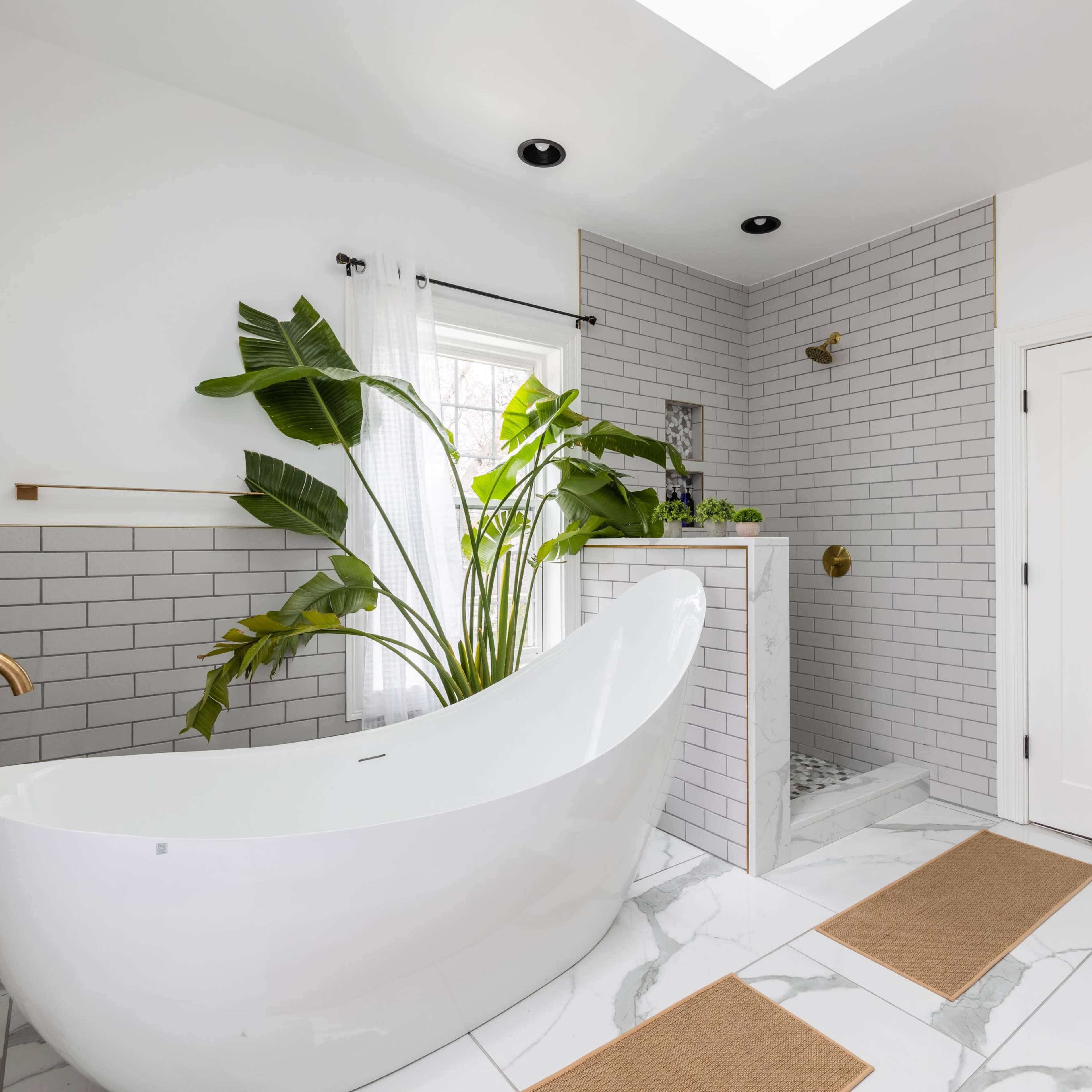 The bathroom features a freestanding white bathtub, a glass-enclosed shower, and large potted plants set against a backdrop of white tile and marble flooring.