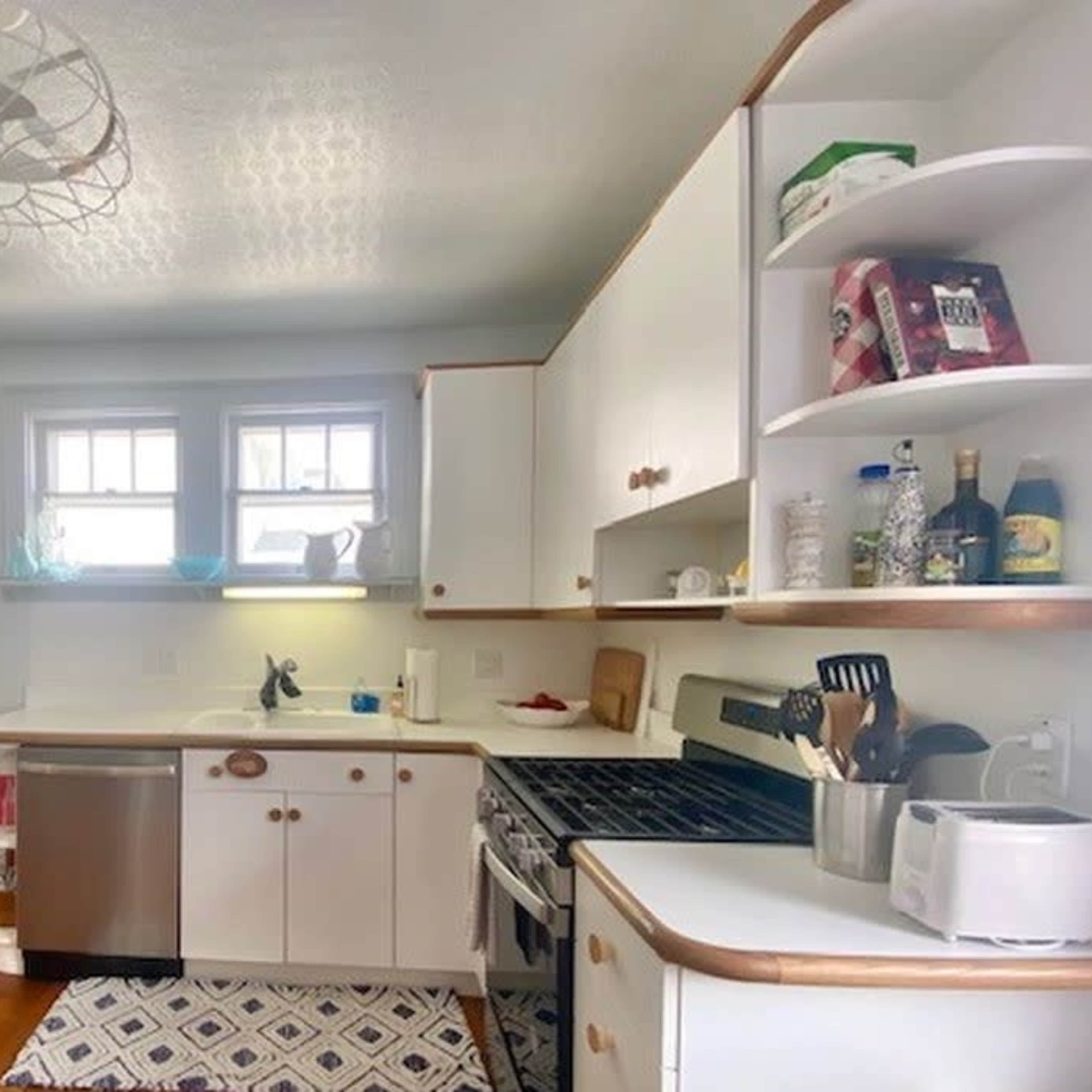 A tidy kitchen features a wooden prep table, stainless steel appliances, and white cabinetry lined with dishes and cooking supplies.
