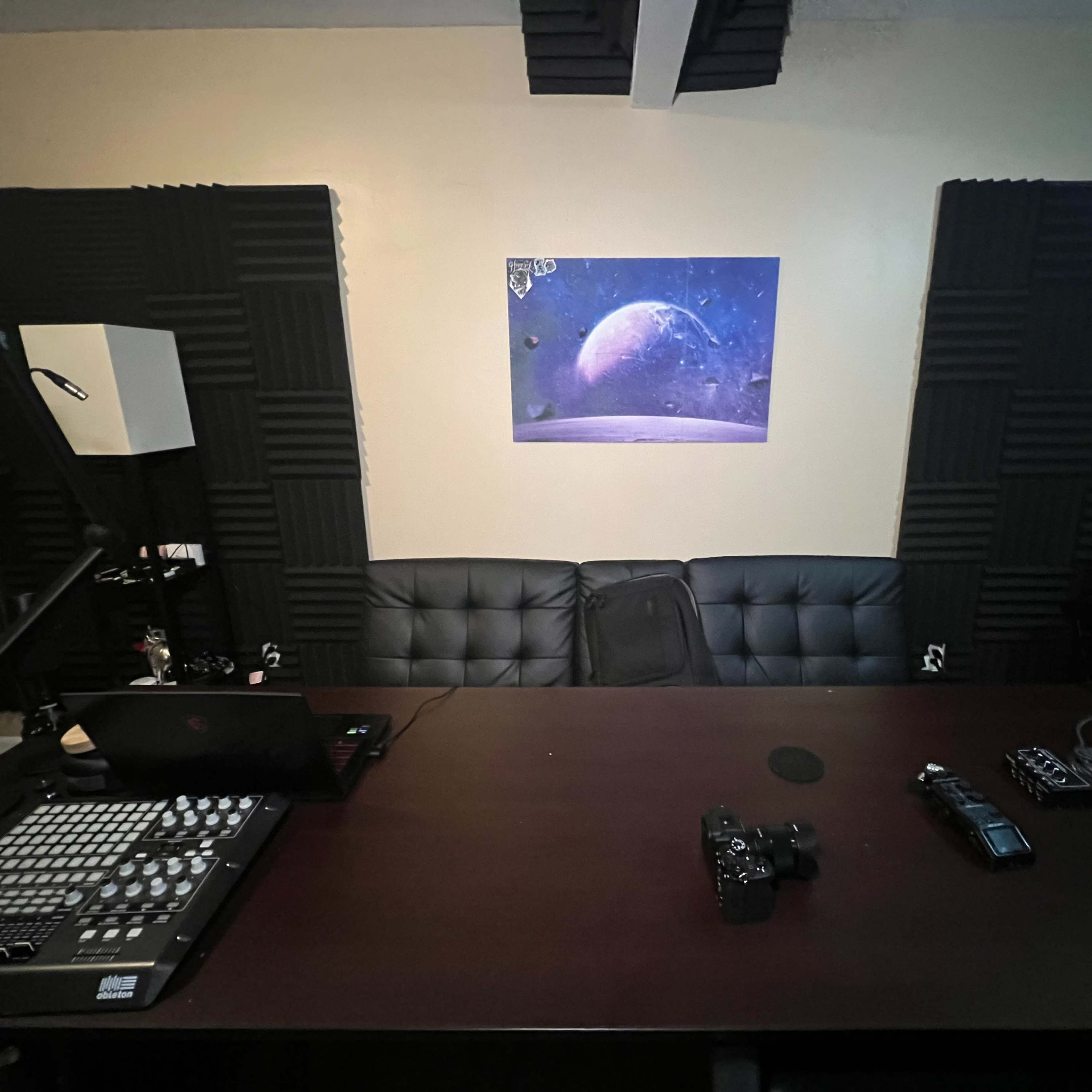 A dimly lit recording studio setup with a desk, audio equipment, and a large print of a planet hanging on the wall.