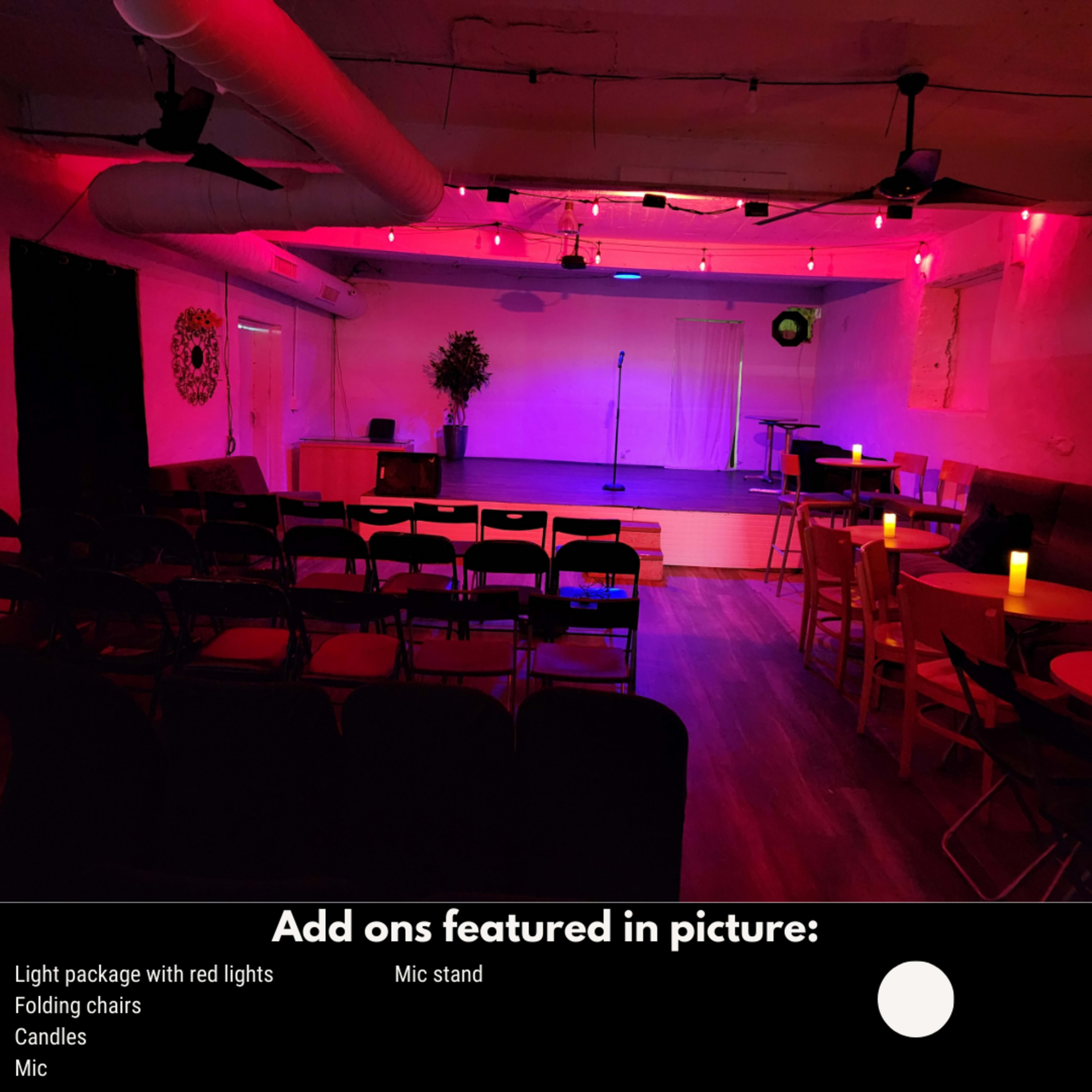 A small performance venue with folding chairs arranged around tables, illuminated by red lights, with a mic stand and candles on the tables.