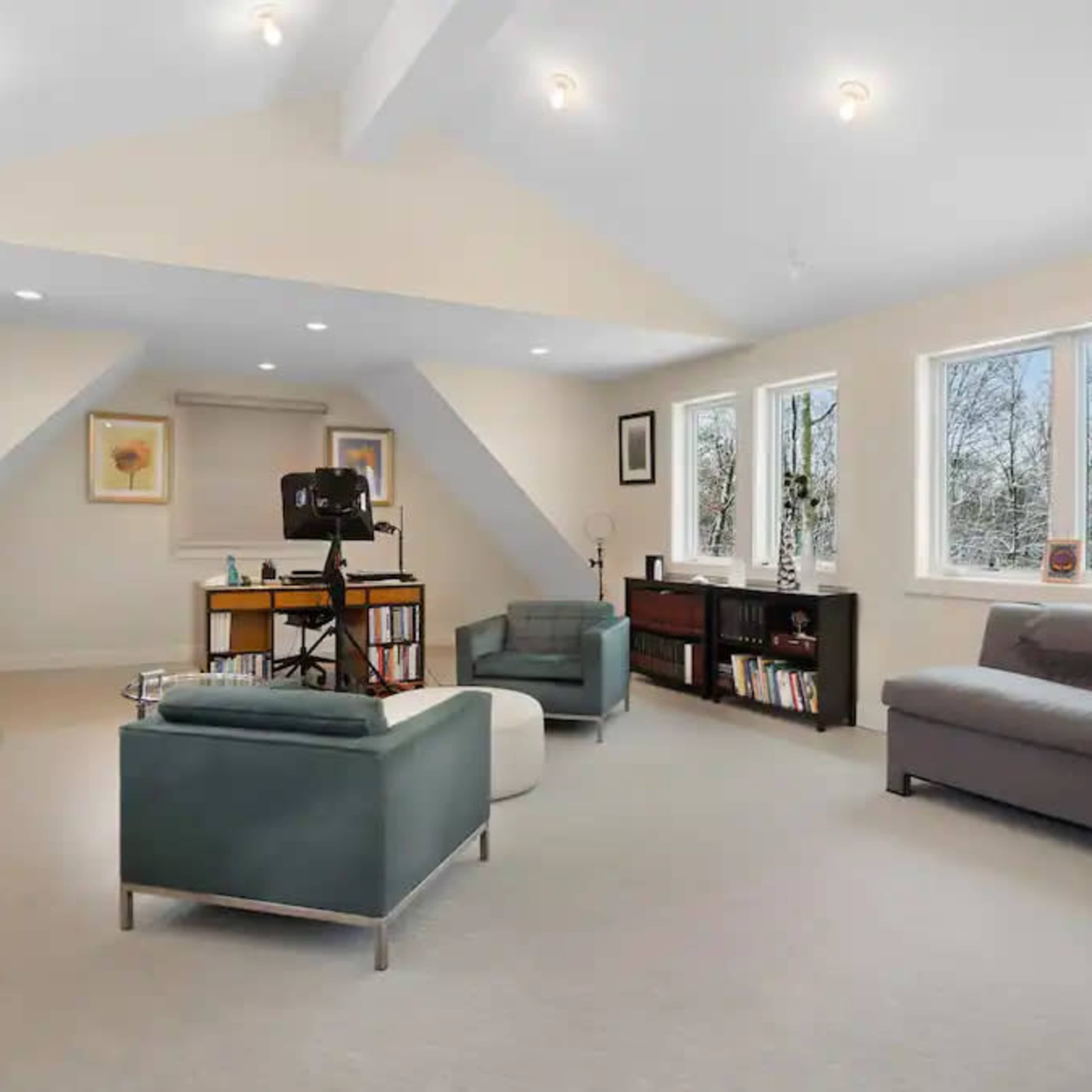 A well-lit attic room with two chairs, a sofa, shelves filled with books, and a desk setup near windows.