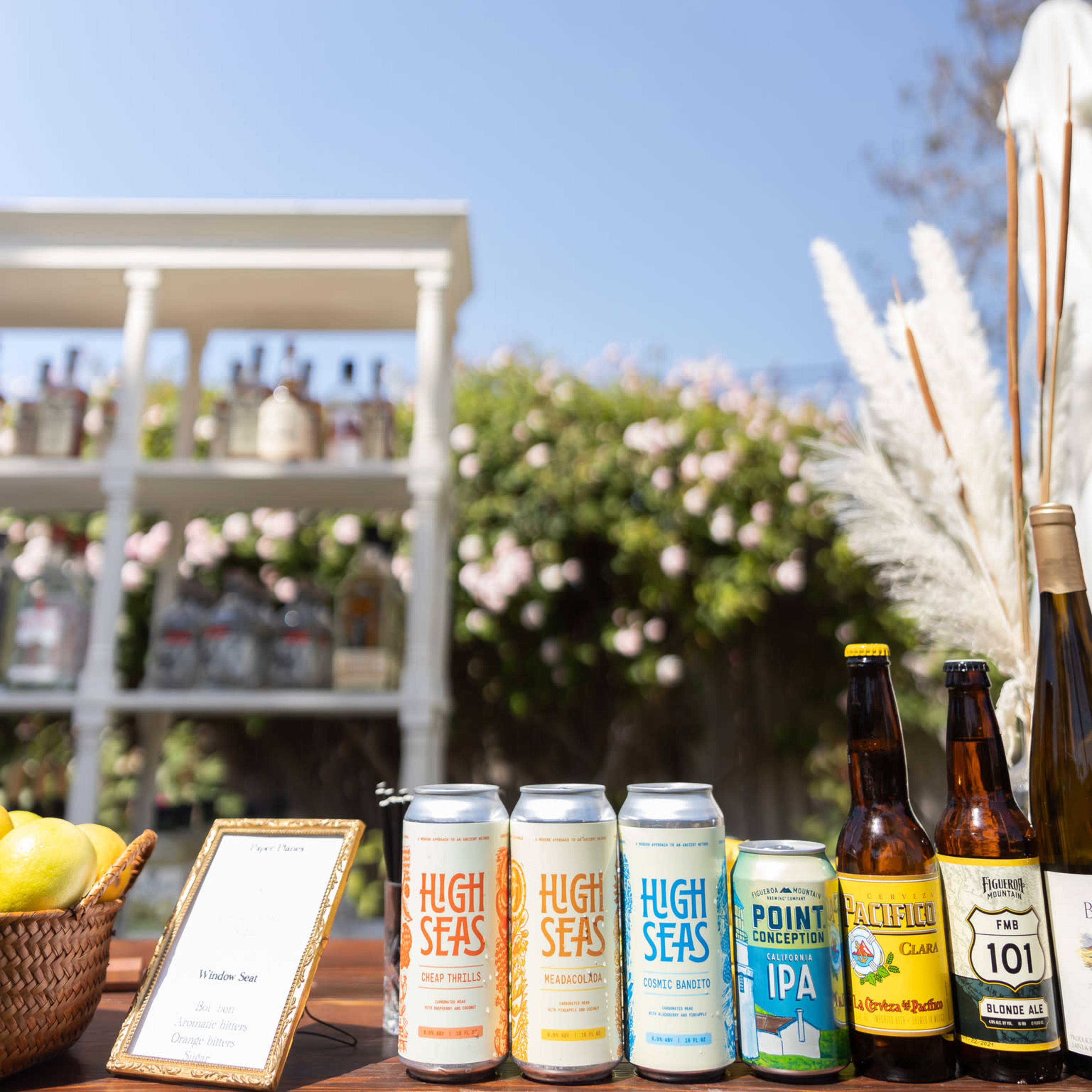 A selection of craft beers is displayed on a table under a clear blue sky, with a background of a shelf holding various bottles.