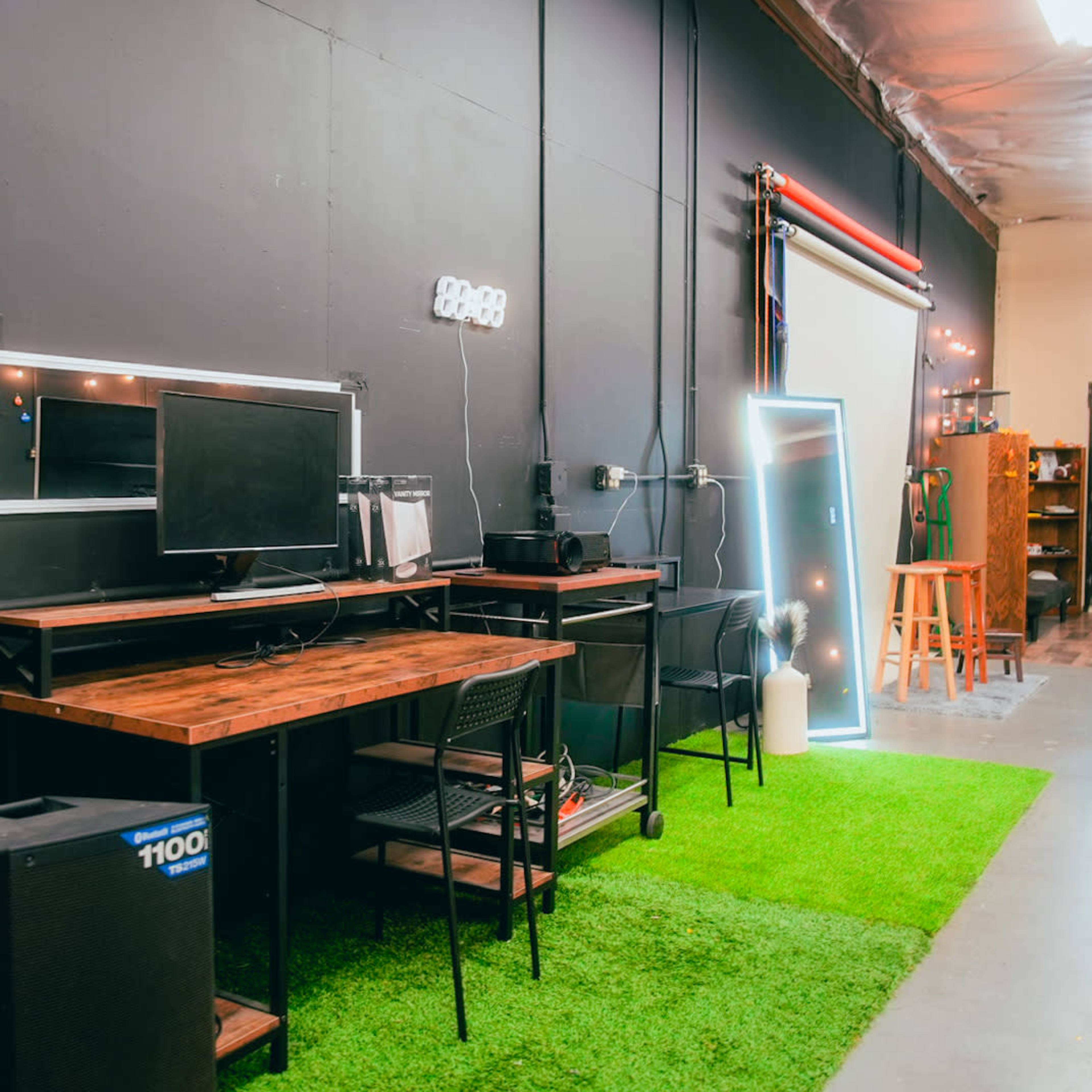 A workspace with a wooden desk, black chair, computer monitor, and a green artificial grass carpet against a dark wall.