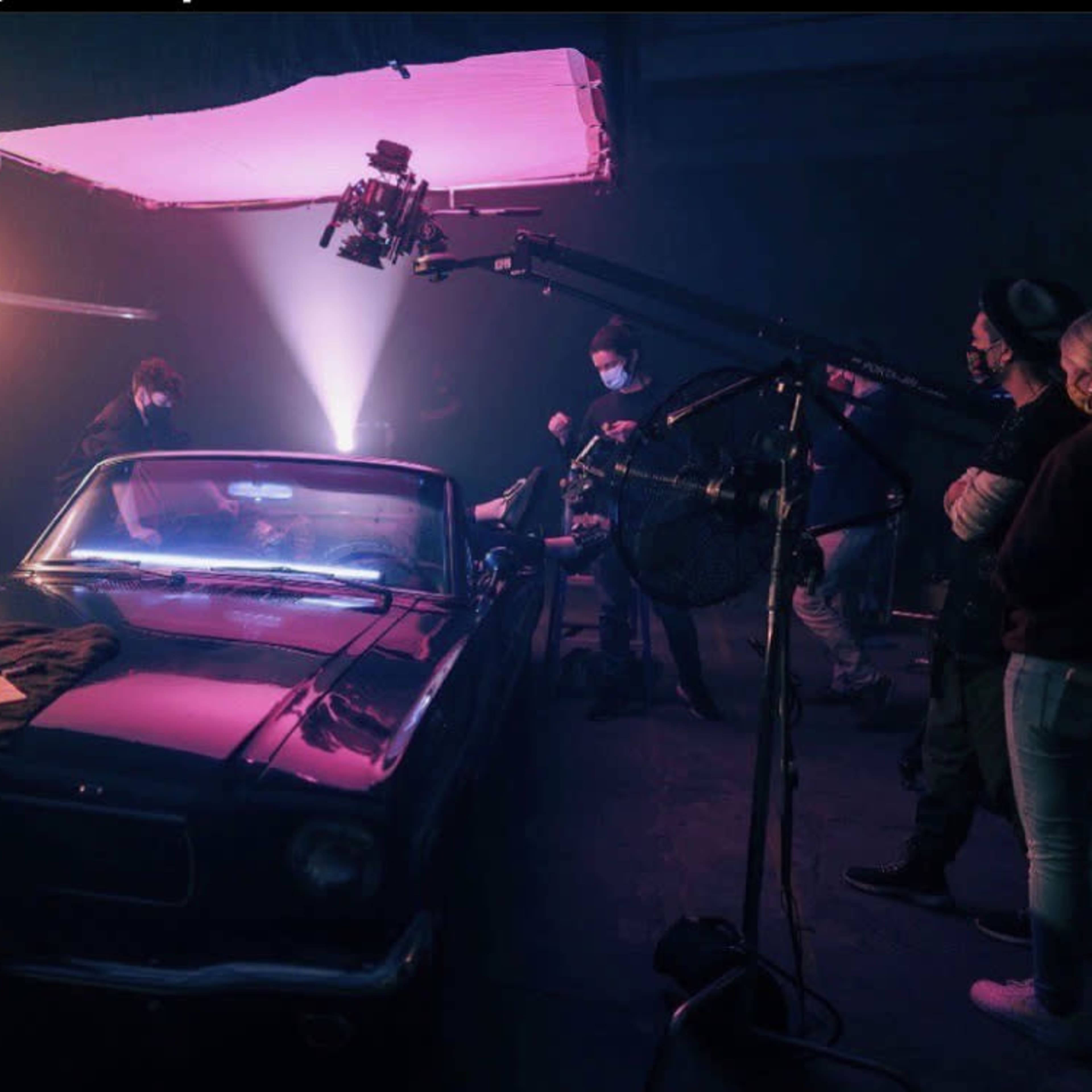 A group of people works around a classic black car in a dimly lit studio, using lighting equipment and cameras for a photoshoot.