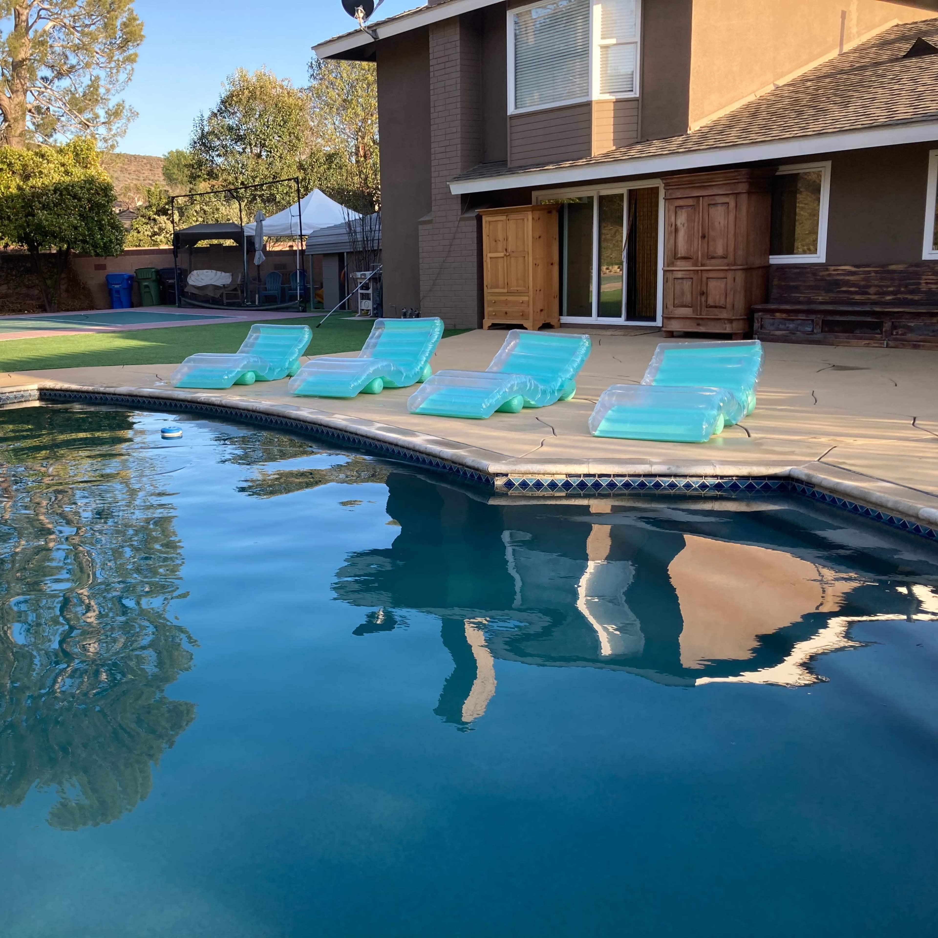 A swimming pool with five turquoise lounge chairs arranged neatly around it, reflecting the surrounding trees and a house with large windows.