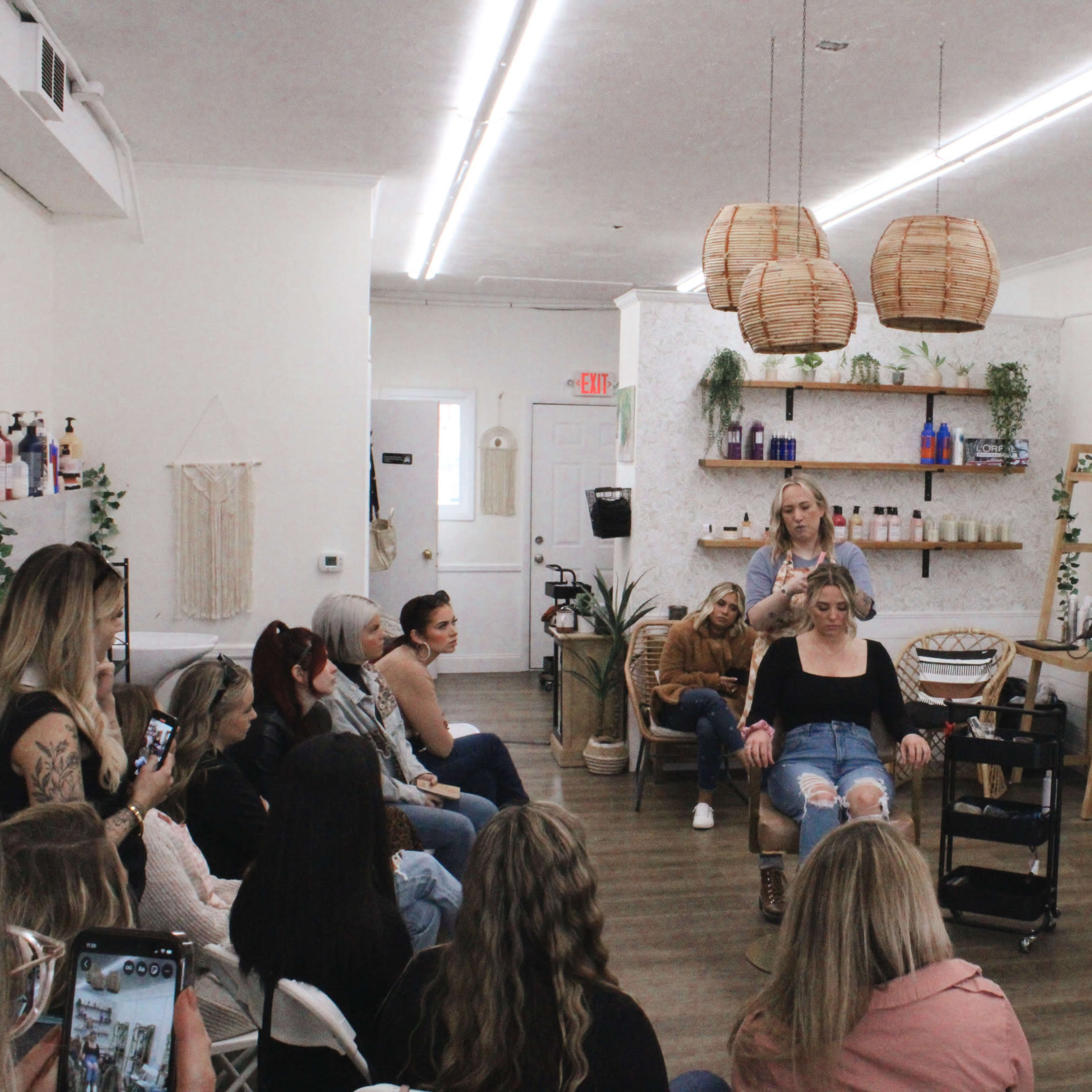 A group of people sits in a salon, watching a stylist demonstrate hair techniques on a model in a chair.