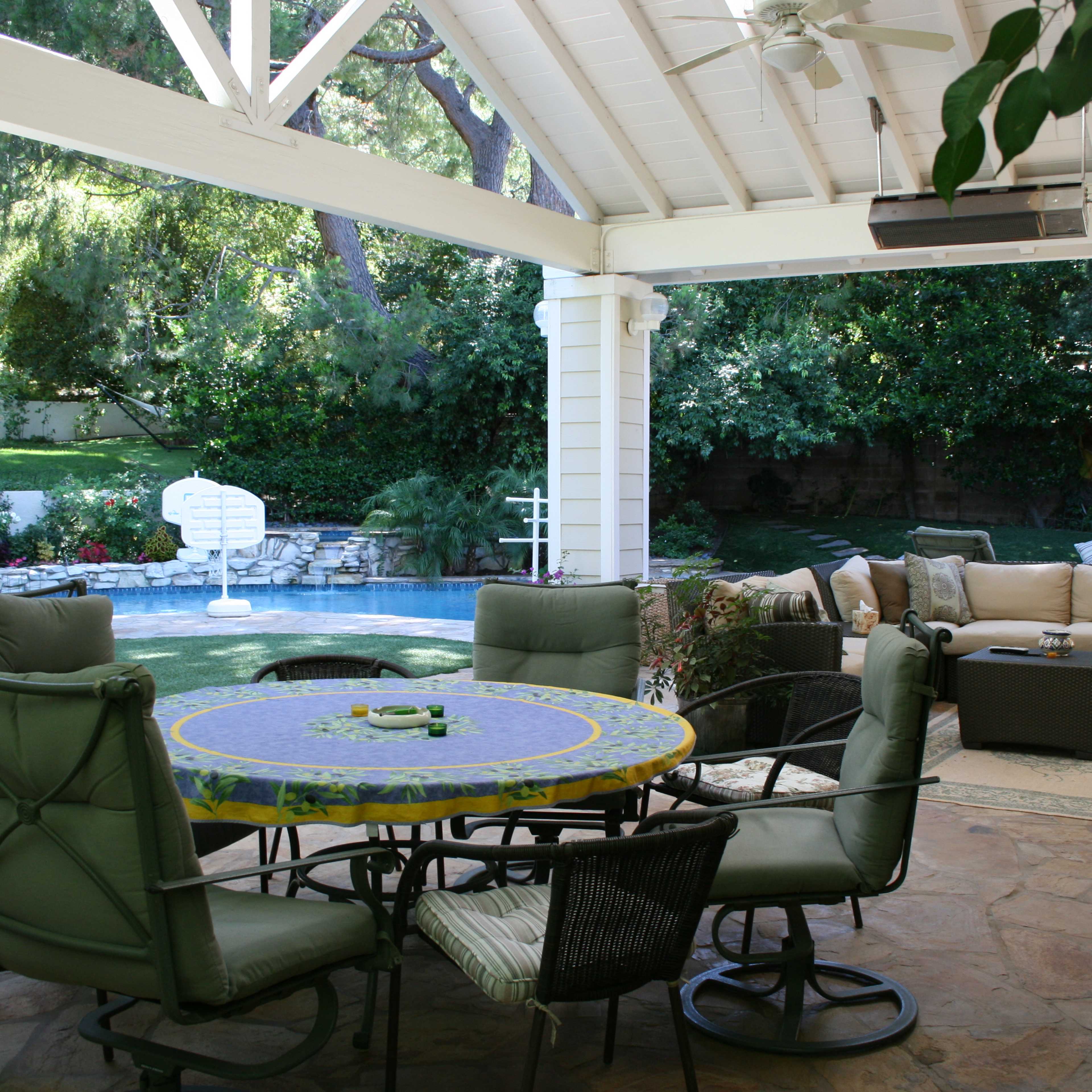 A backyard patio with a round dining table, green chairs, and a seating area beside a pool surrounded by greenery.