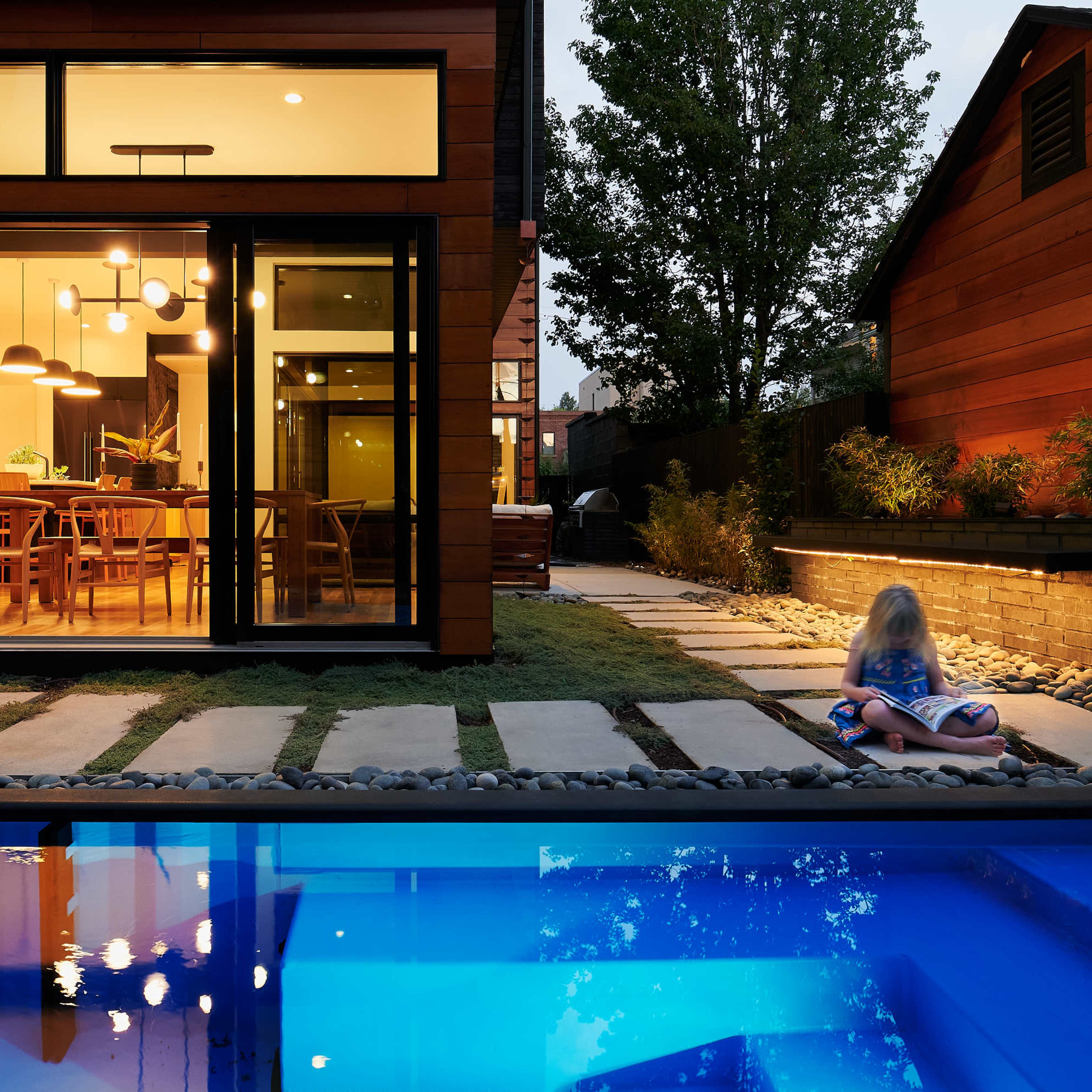 A child sits on a towel near a swimming pool in a backyard, with a modern house featuring large windows in the background.