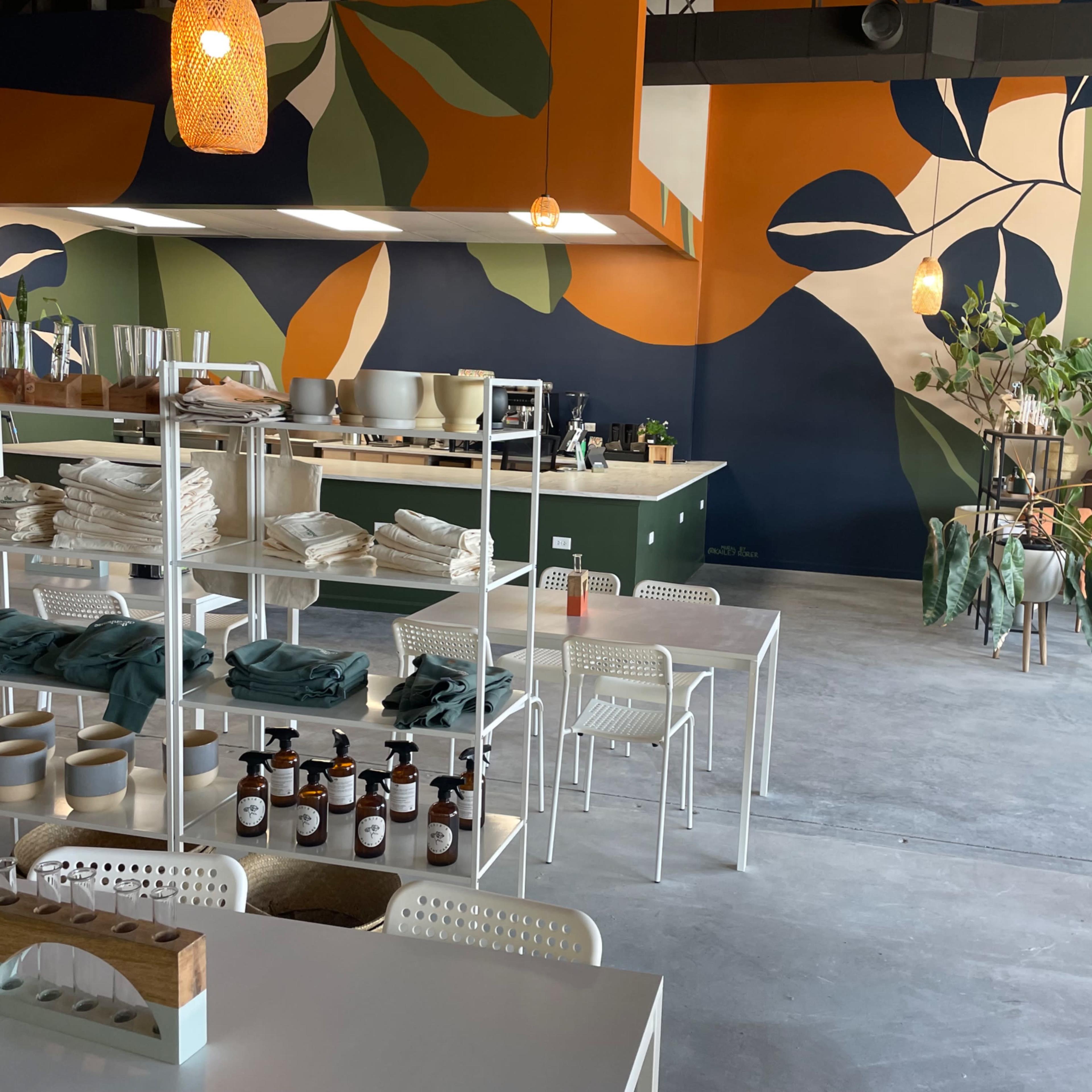The interior of a café features a colorful mural, white furniture, and shelves displaying neatly arranged items and plants.