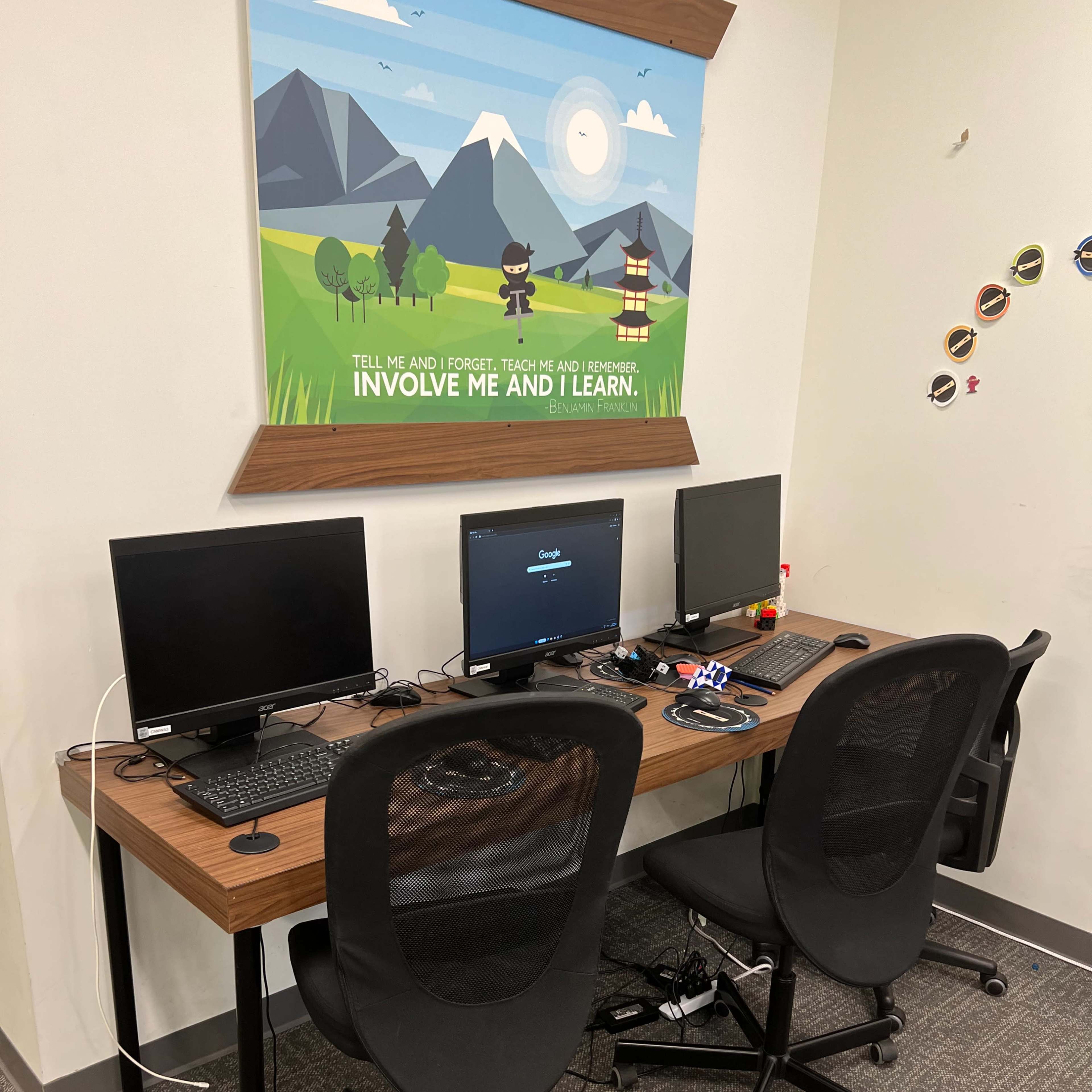 A workspace features two computer desks with monitors, located against a wall adorned with a decorative poster.