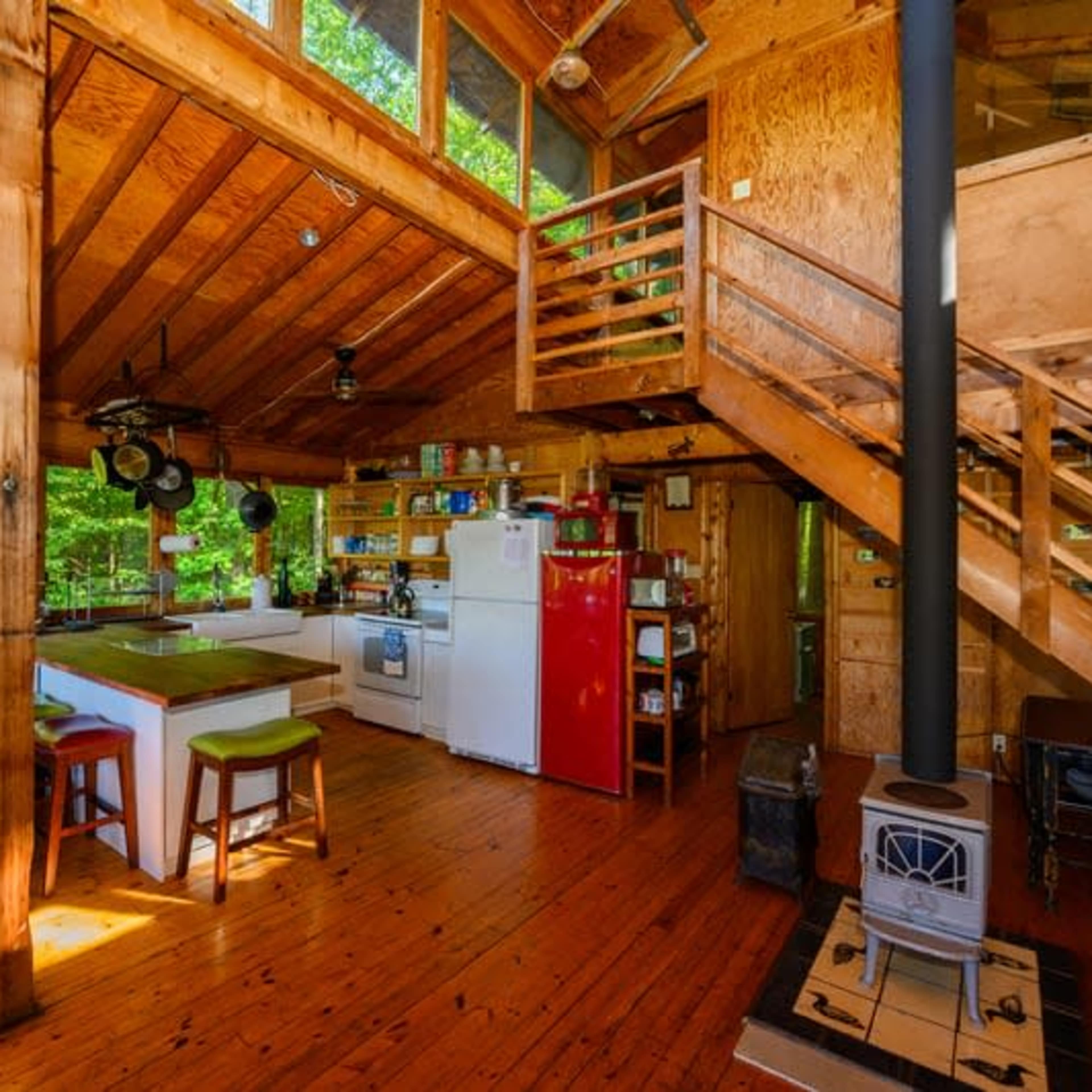 The interior of a wooden cabin with an open kitchen, a staircase, and a cozy living area featuring a stove.
