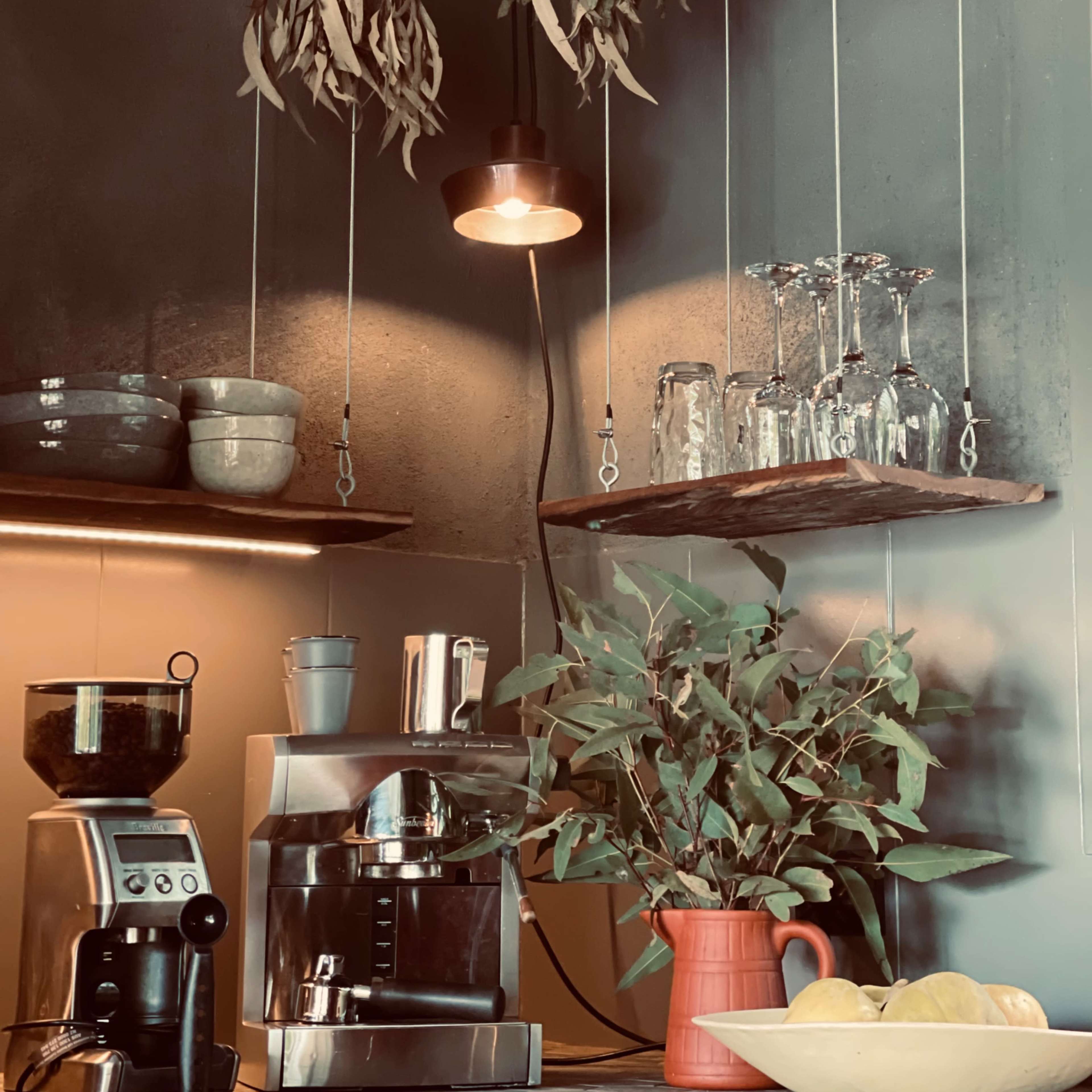 A coffee preparation area with hanging dried leaves, a suspended light fixture, a coffee machine, a grinder, and a shelf holding glasses and pottery.