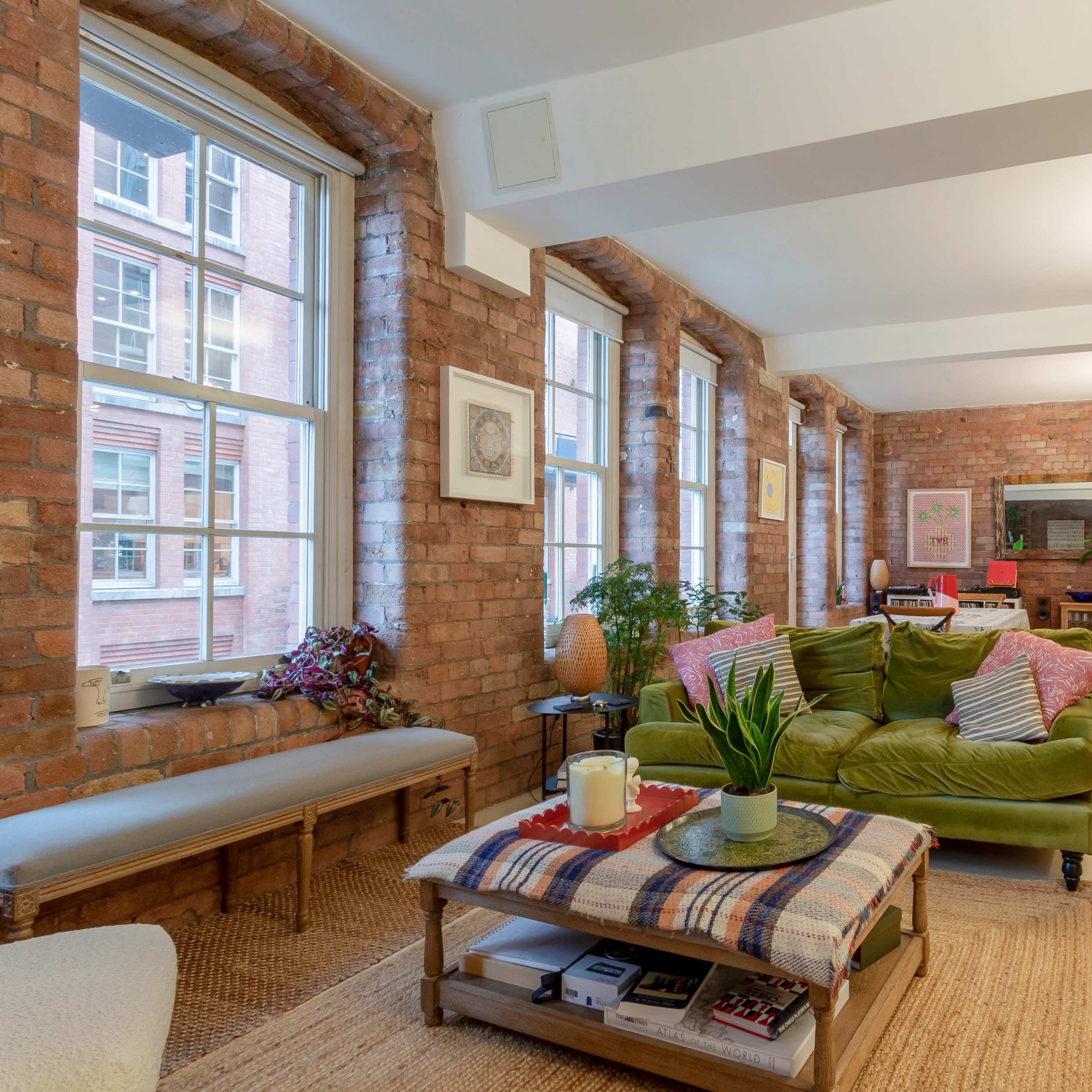 A modern living space with large windows, exposed brick walls, a green sofa, and a wooden coffee table adorned with a plant and a candle.