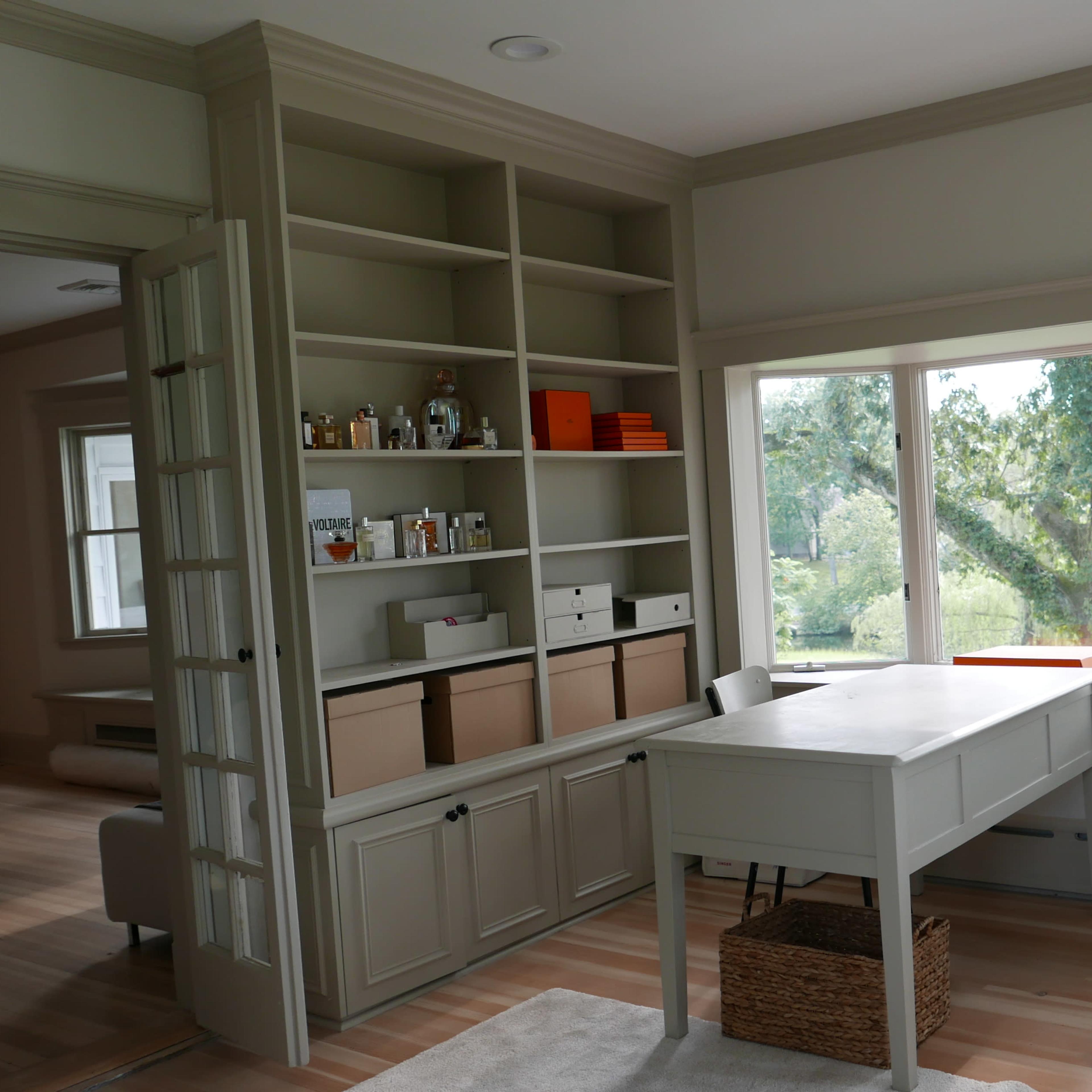 A neatly organized room featuring a light-colored desk, shelving with various boxes and jars, and a large window overlooking a green landscape.