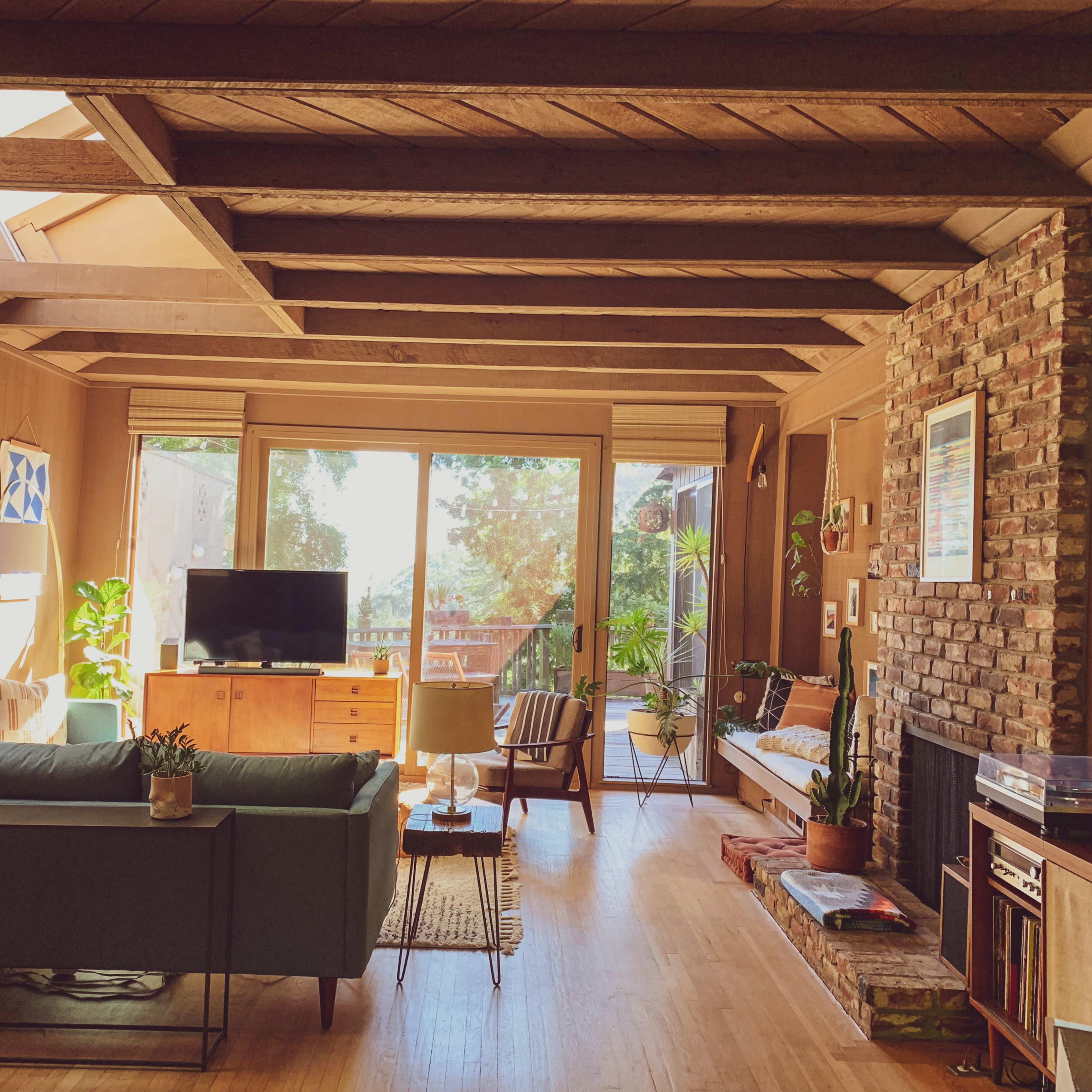 A cozy living room with wooden beams, a large window overlooking greenery, a sectional sofa, a television, and a stone fireplace.