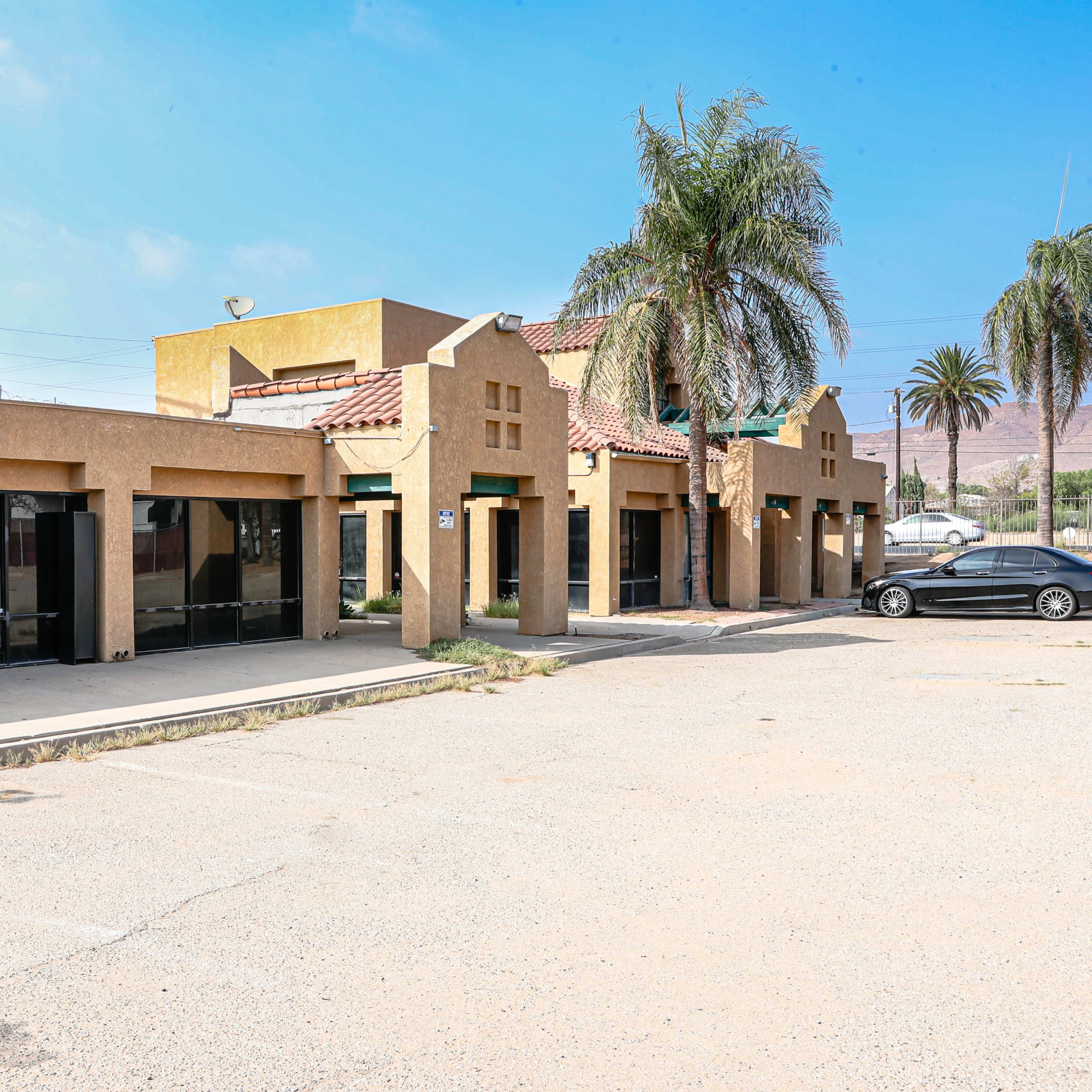 A row of single-story commercial buildings with a stucco facade and tile roofs sits adjacent to a parking lot lined with palm trees.