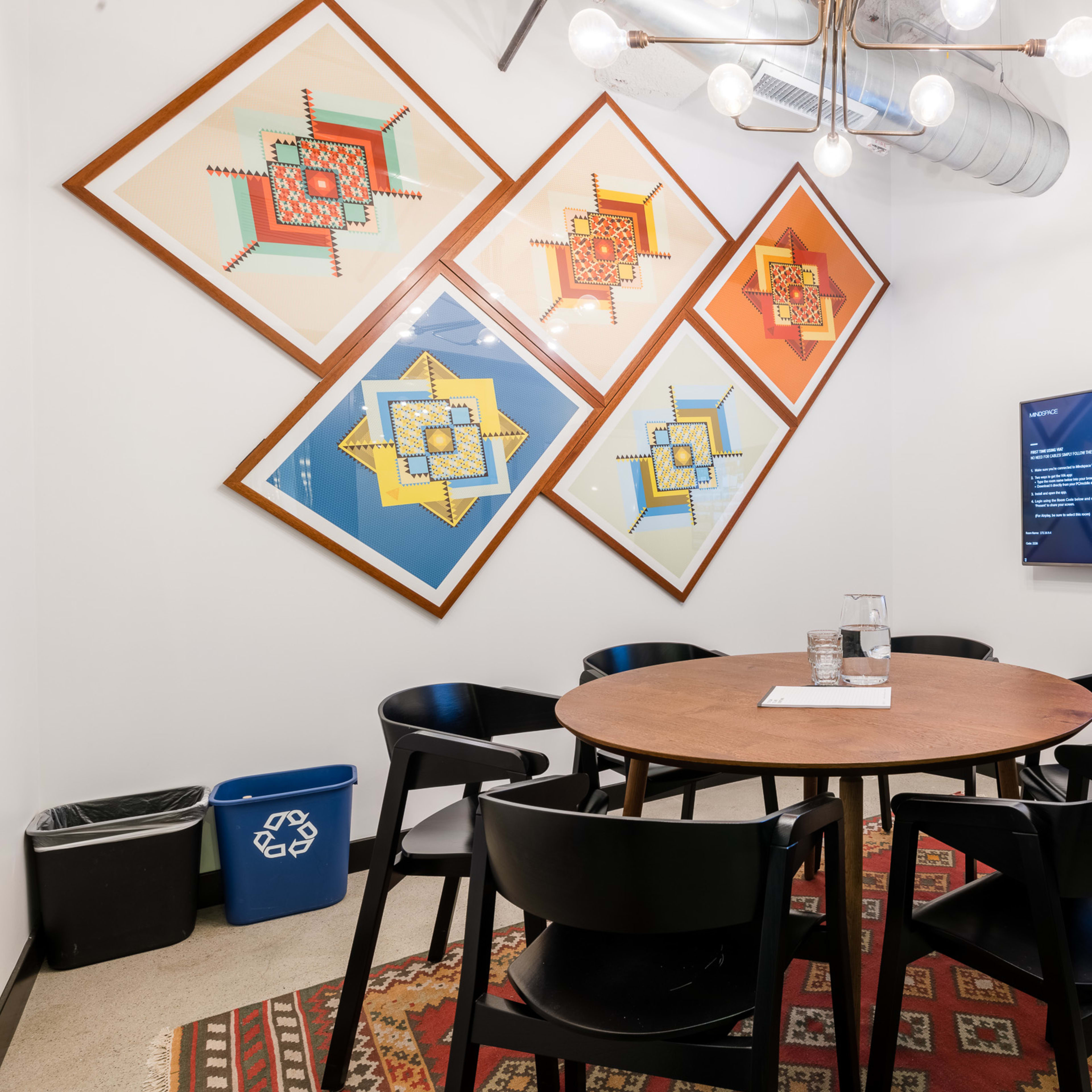 A conference room features a round wooden table surrounded by black chairs, with colorful wall art and a television displaying information.