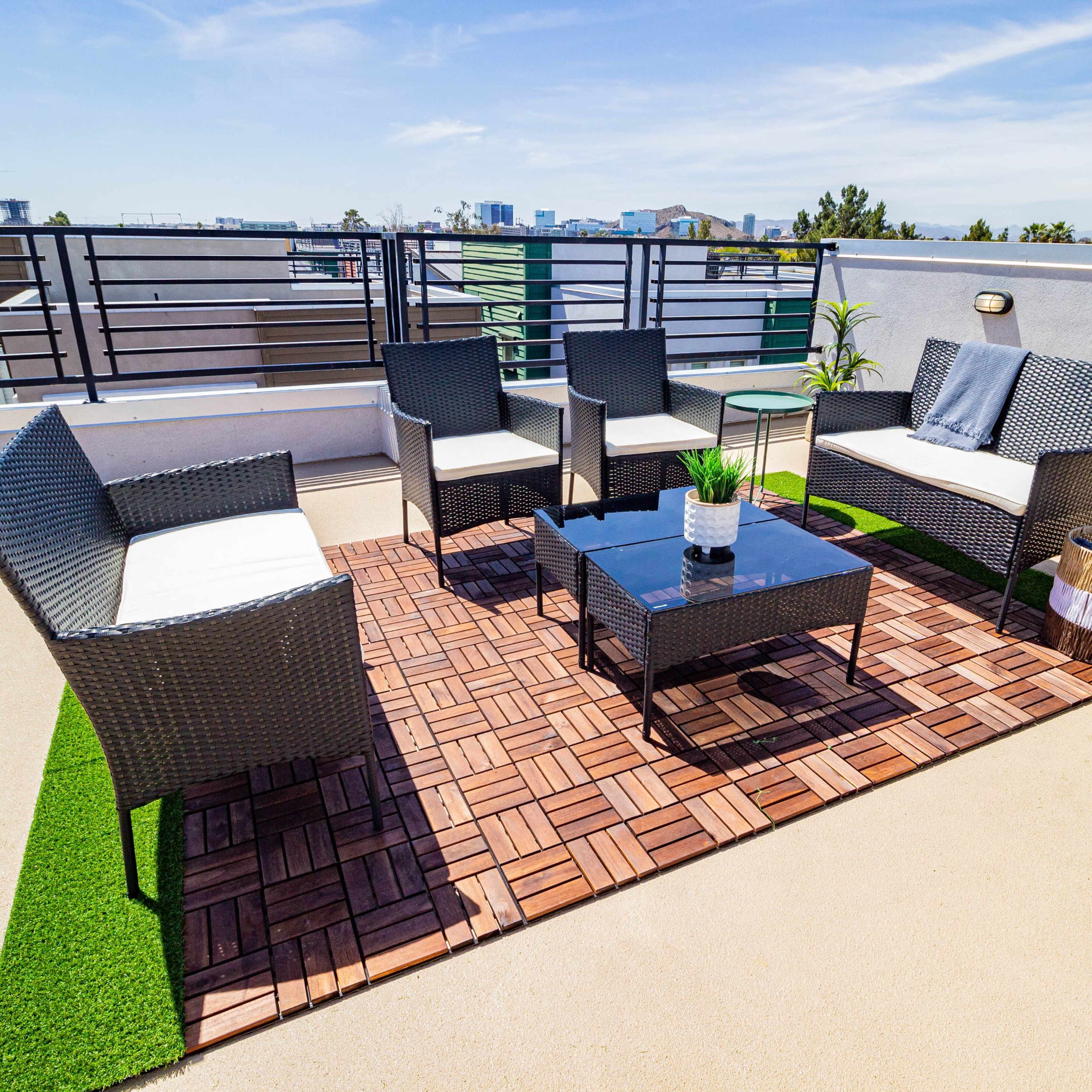 A rooftop seating area with four wicker chairs, a glass coffee table, and a decorative plant on a wooden and grass-like floor.