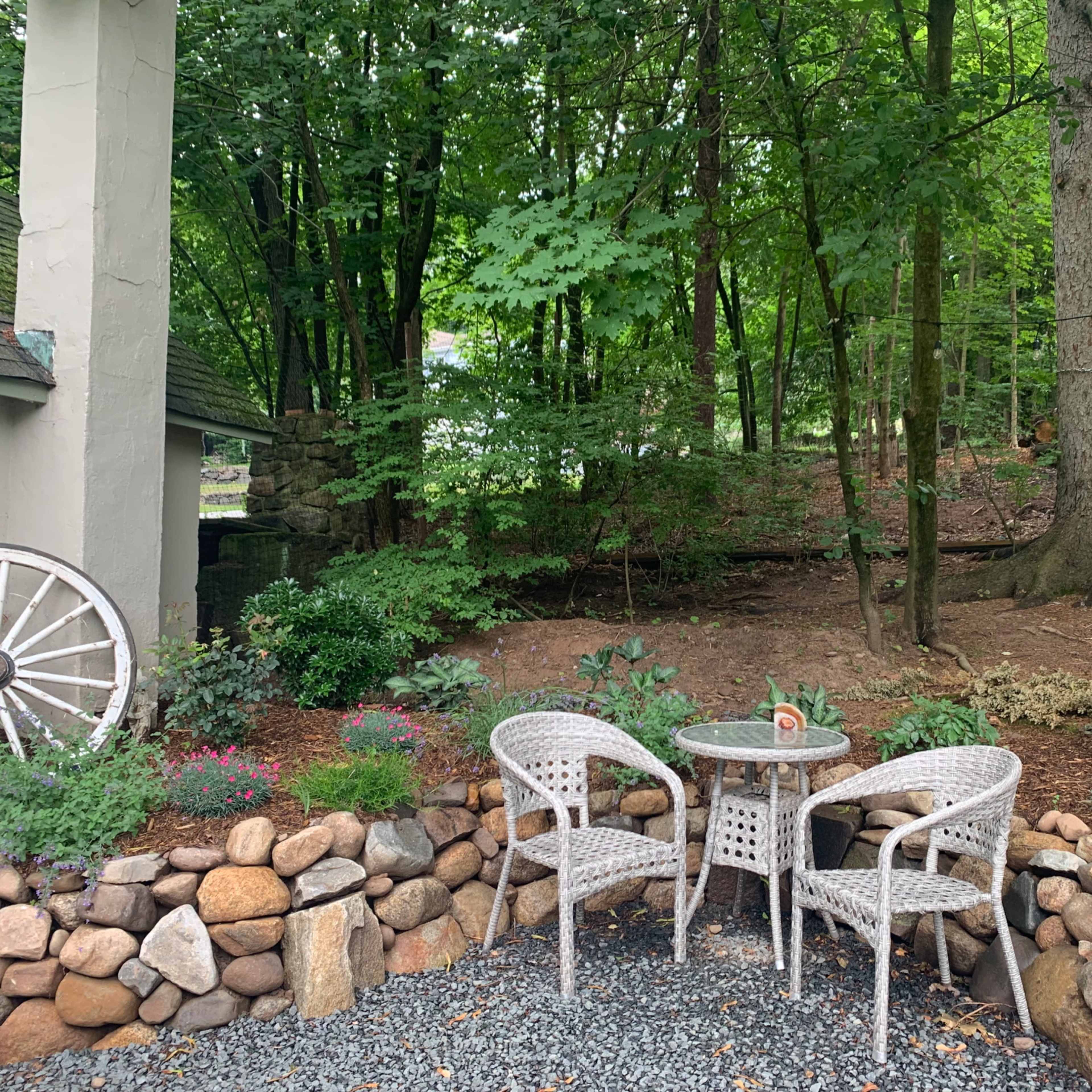 A small outdoor seating area with two white chairs and a table is surrounded by a stone border and lush greenery.