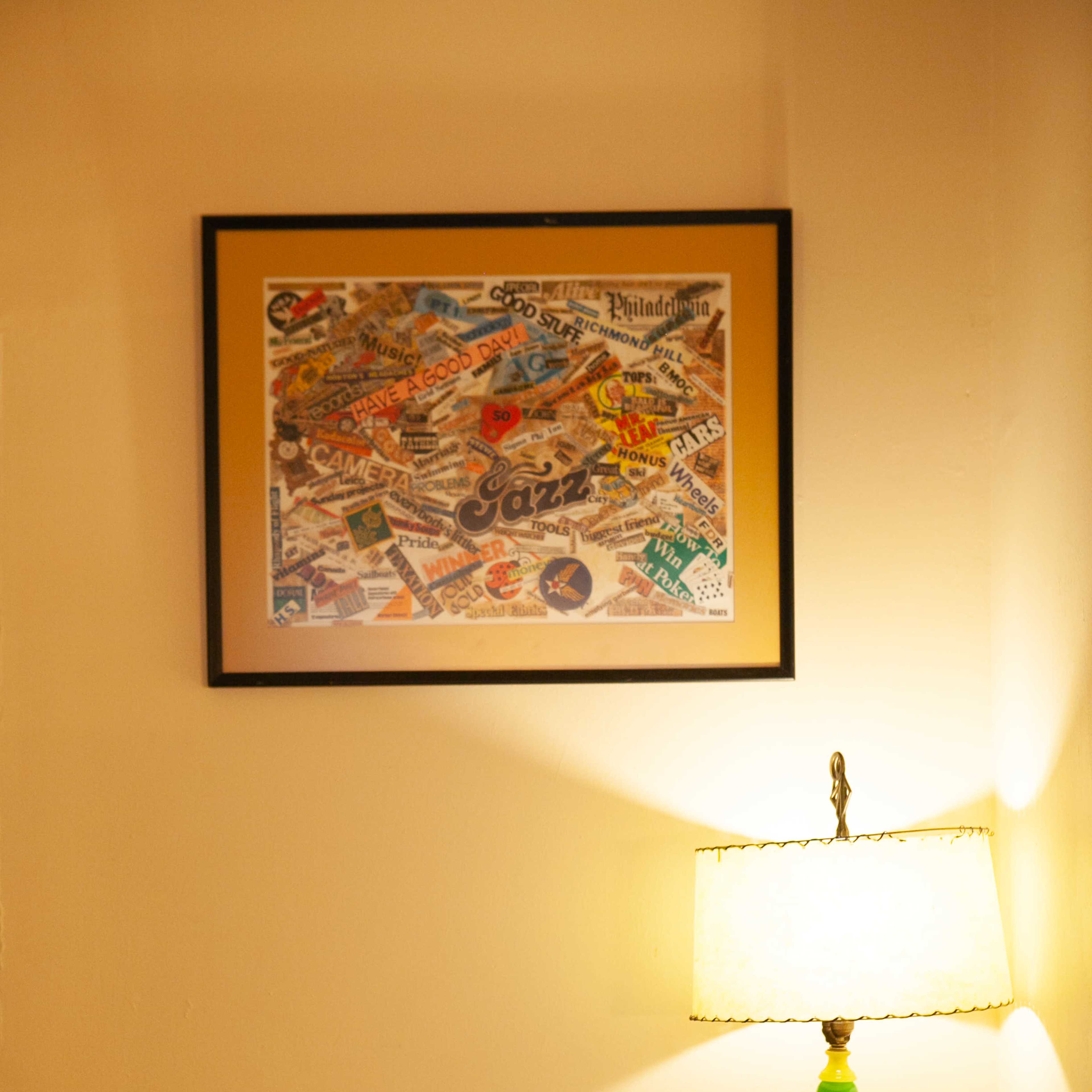 A desk is positioned against a wall, featuring a framed collage of newspaper clippings above it, a lamp to the right, and an empty picture frame on the desk.