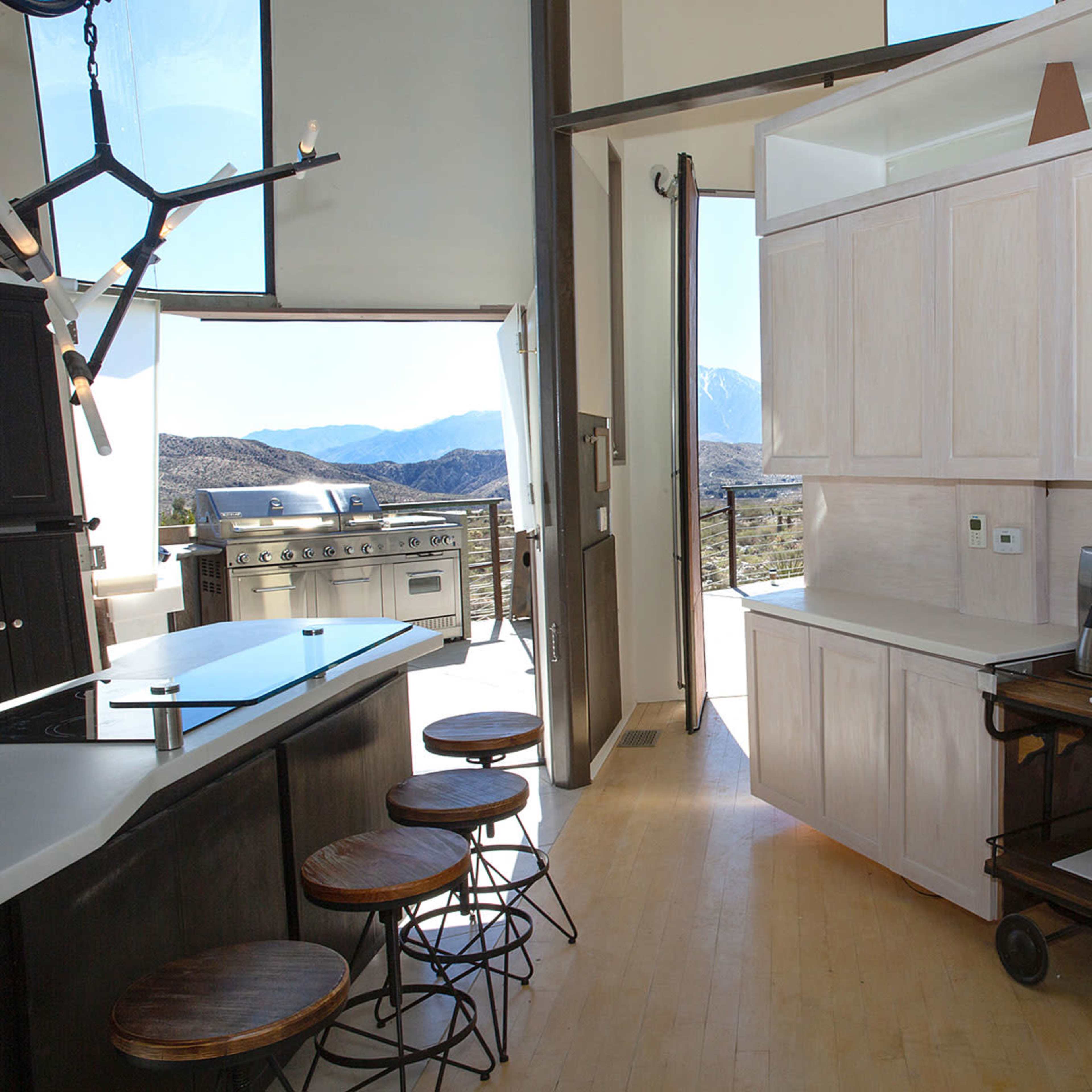 A modern kitchen space featuring a central island with stools, a coffee station on a cart, and large windows overlooking a mountainous landscape.