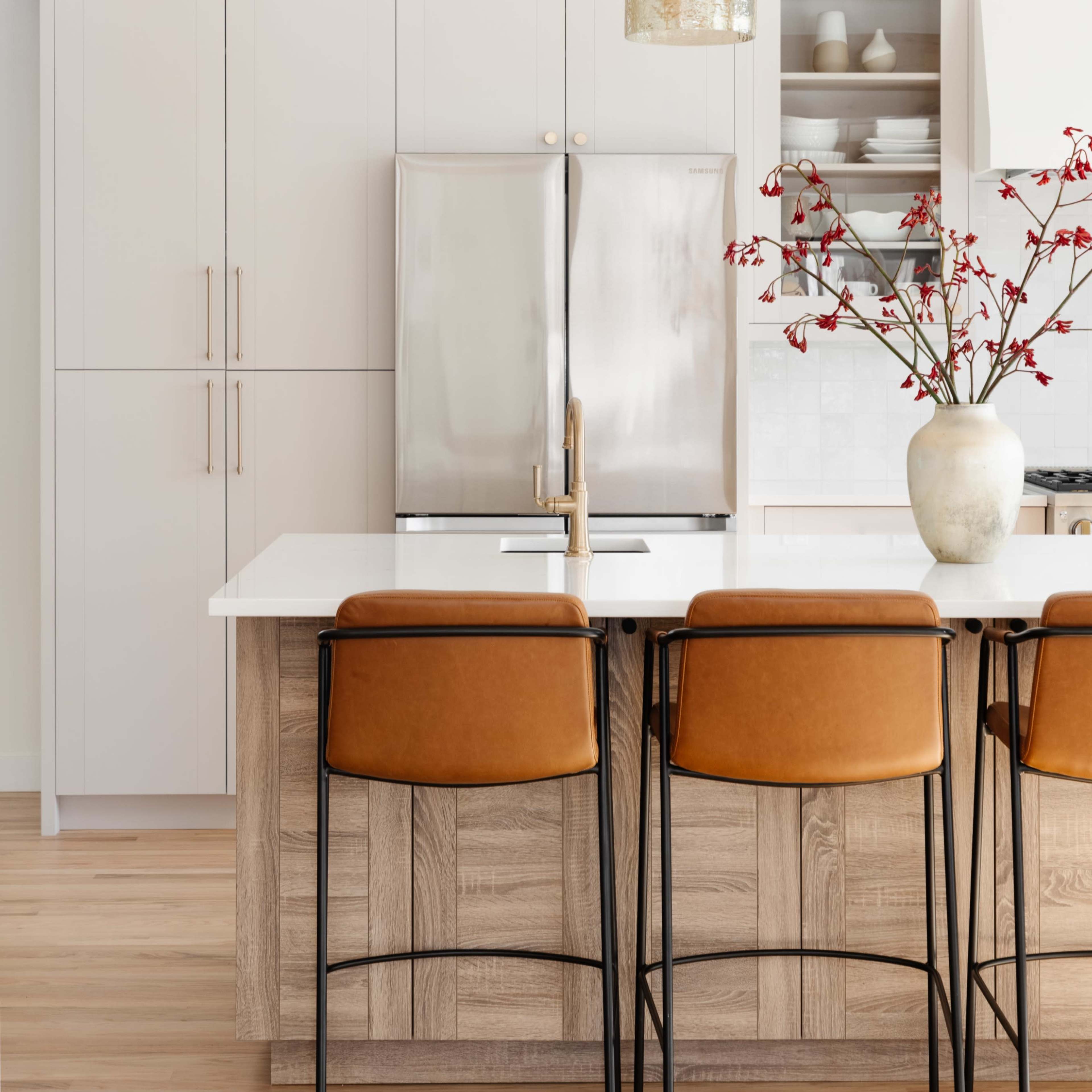A modern kitchen features a large island with leather stools, a stainless steel refrigerator, and a decorative vase with branches.