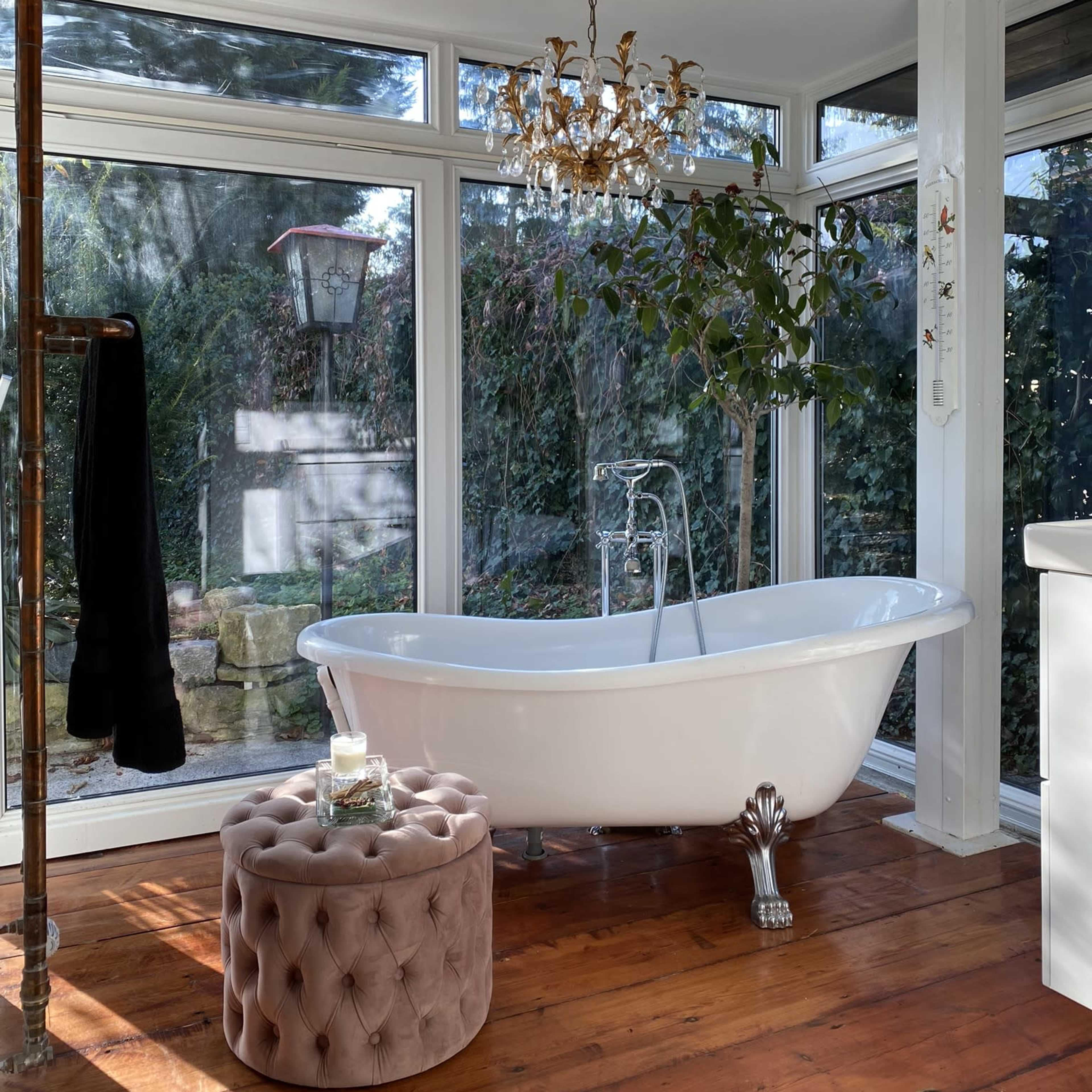 The bathroom features a freestanding white bathtub next to a plush pink ottoman, with large windows providing views of the greenery outside.