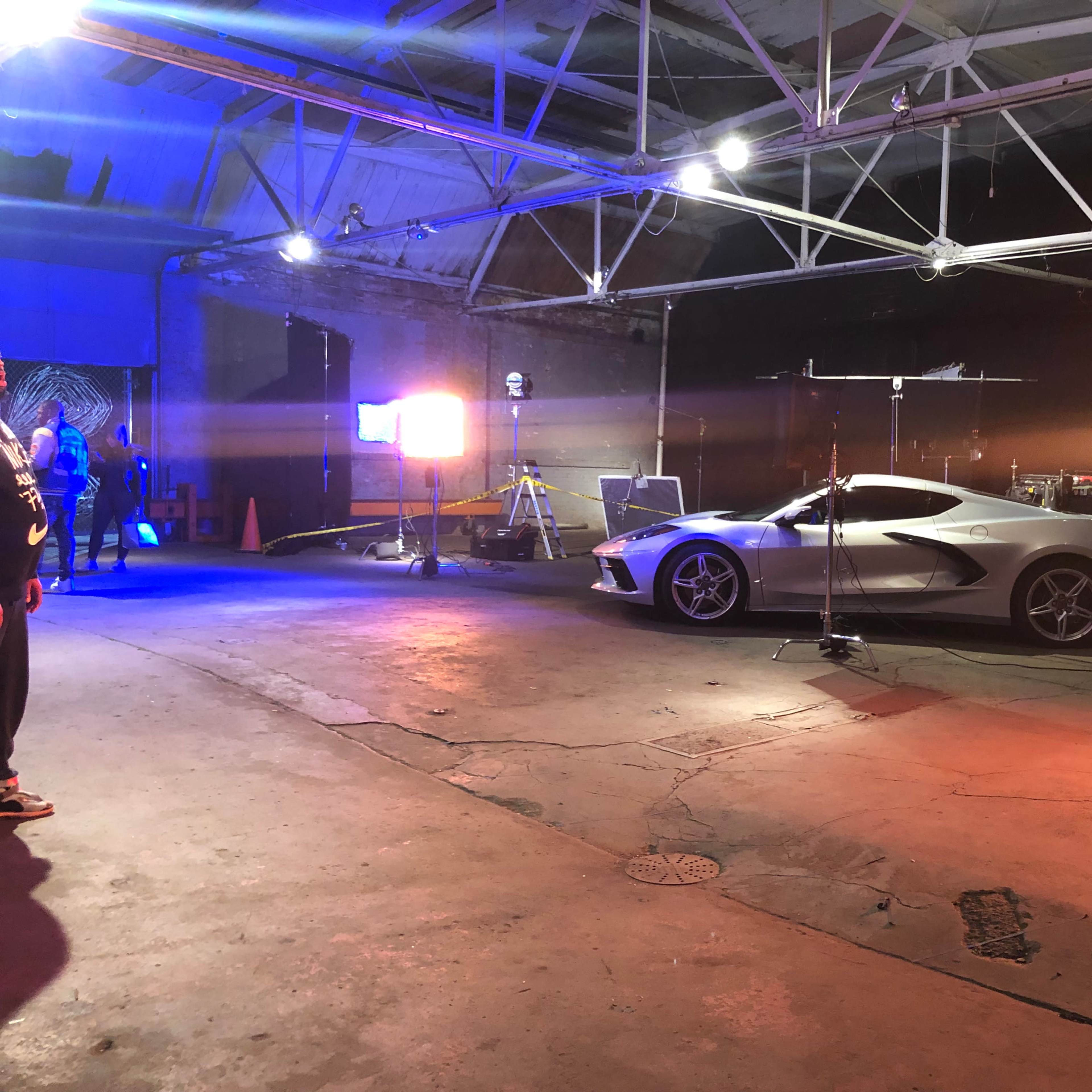 A sleek silver sports car is parked in a well-lit studio space with equipment and a drum set visible in the background.