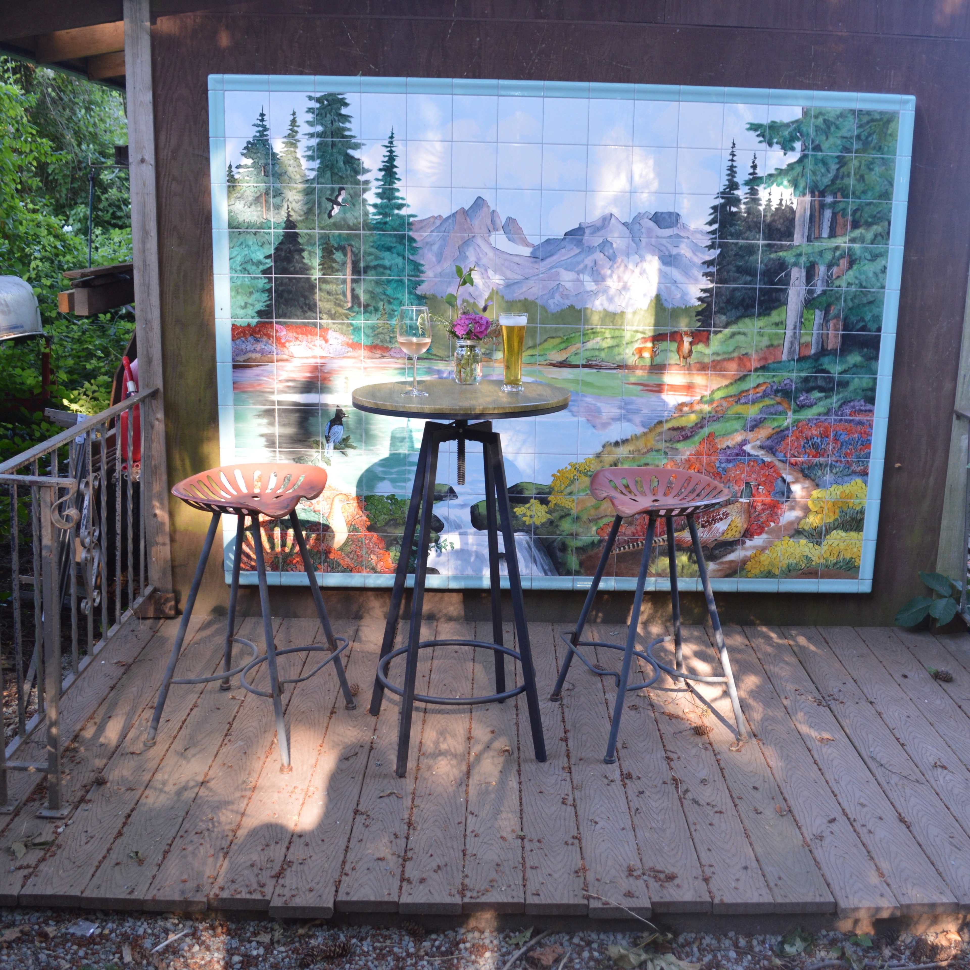 A small wooden deck features two high stools and a table, adjacent to a large mural depicting a scenic landscape with mountains and trees.