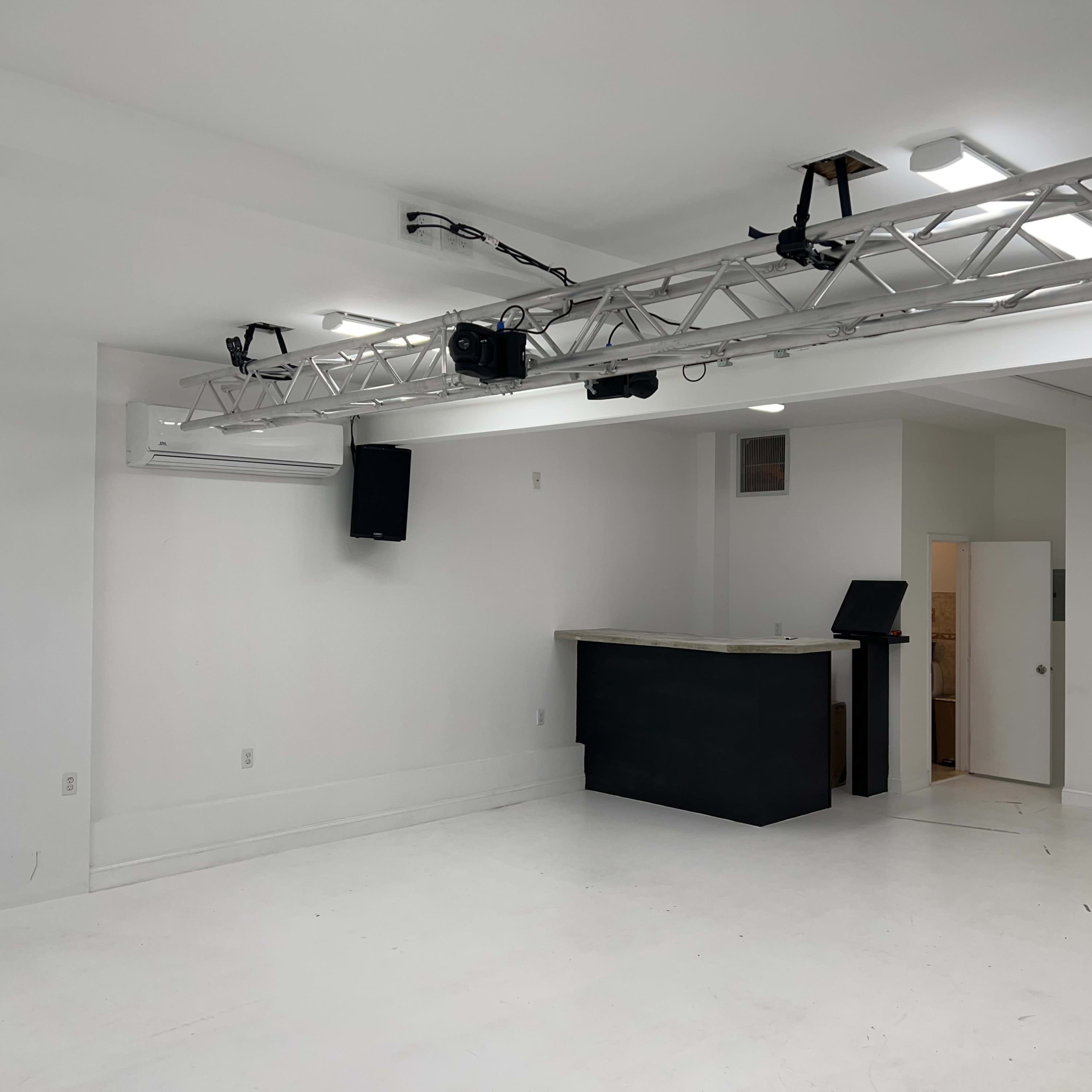 A spacious, minimalistic room with white walls and a large open area, featuring a bar counter and a lighting truss overhead.