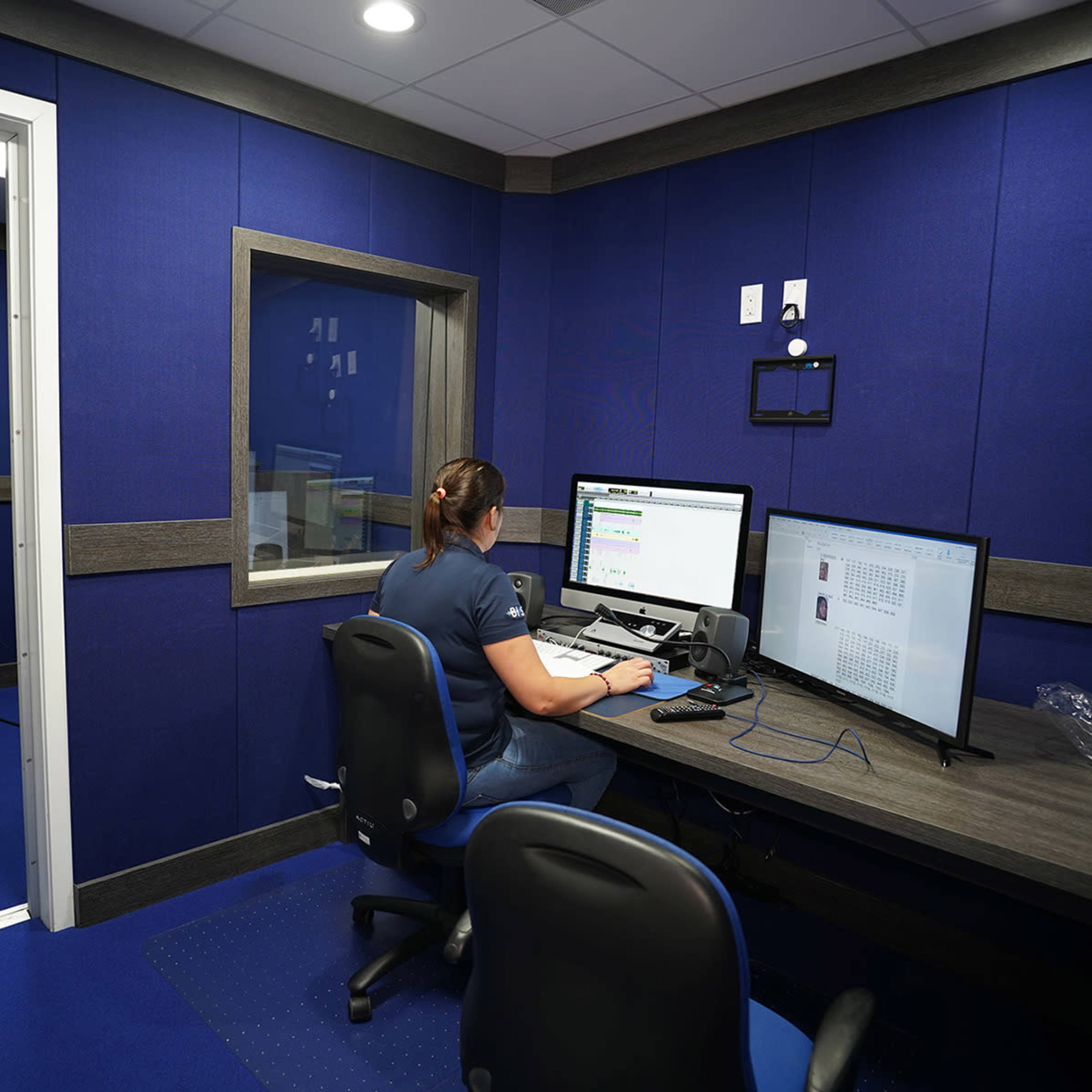 A person is working at a desk with dual monitors in a room featuring blue walls and a small window.
