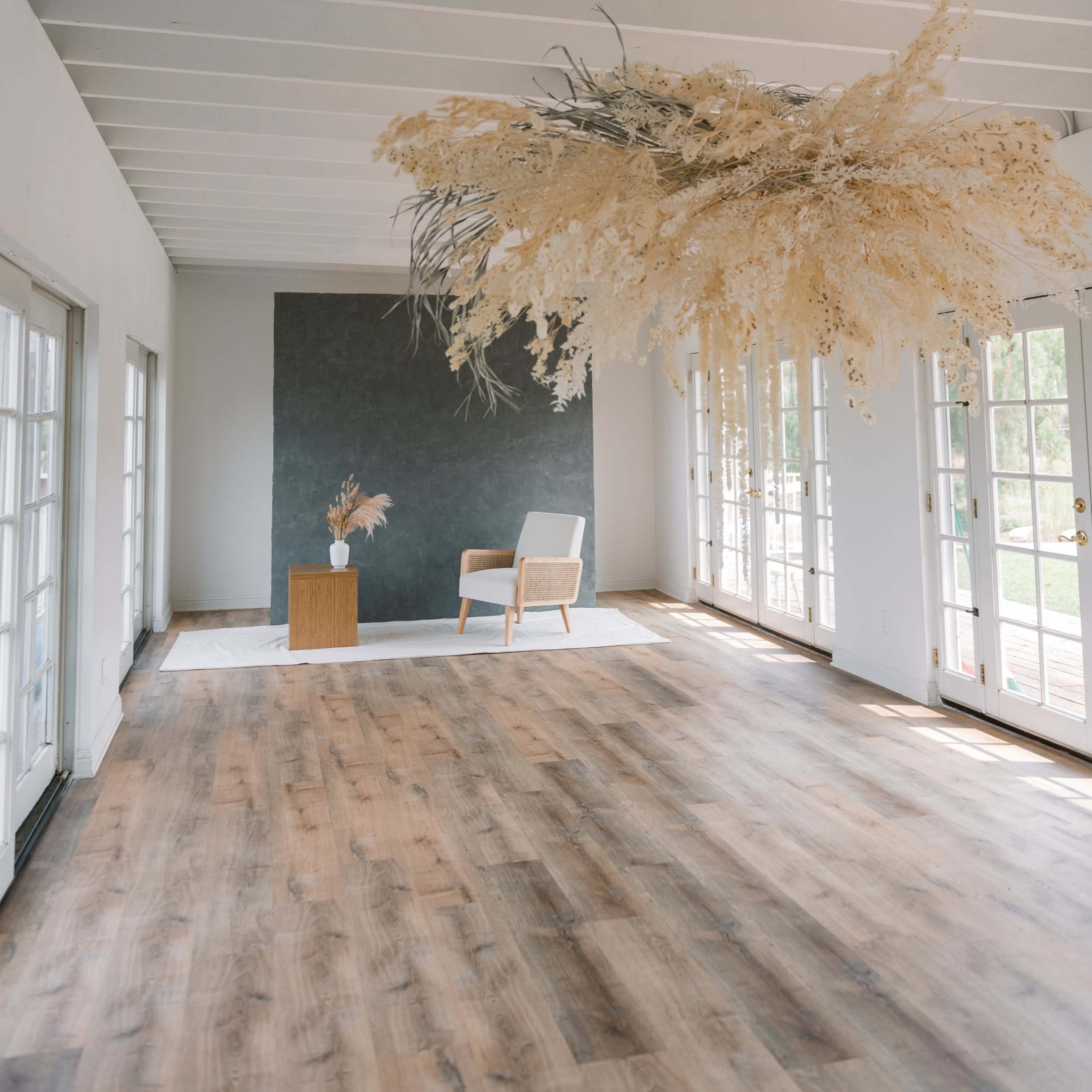 A minimalist interior room with wooden floors, large windows, and a striking dried floral arrangement suspended from the ceiling above a simple wooden side table and an armchair.