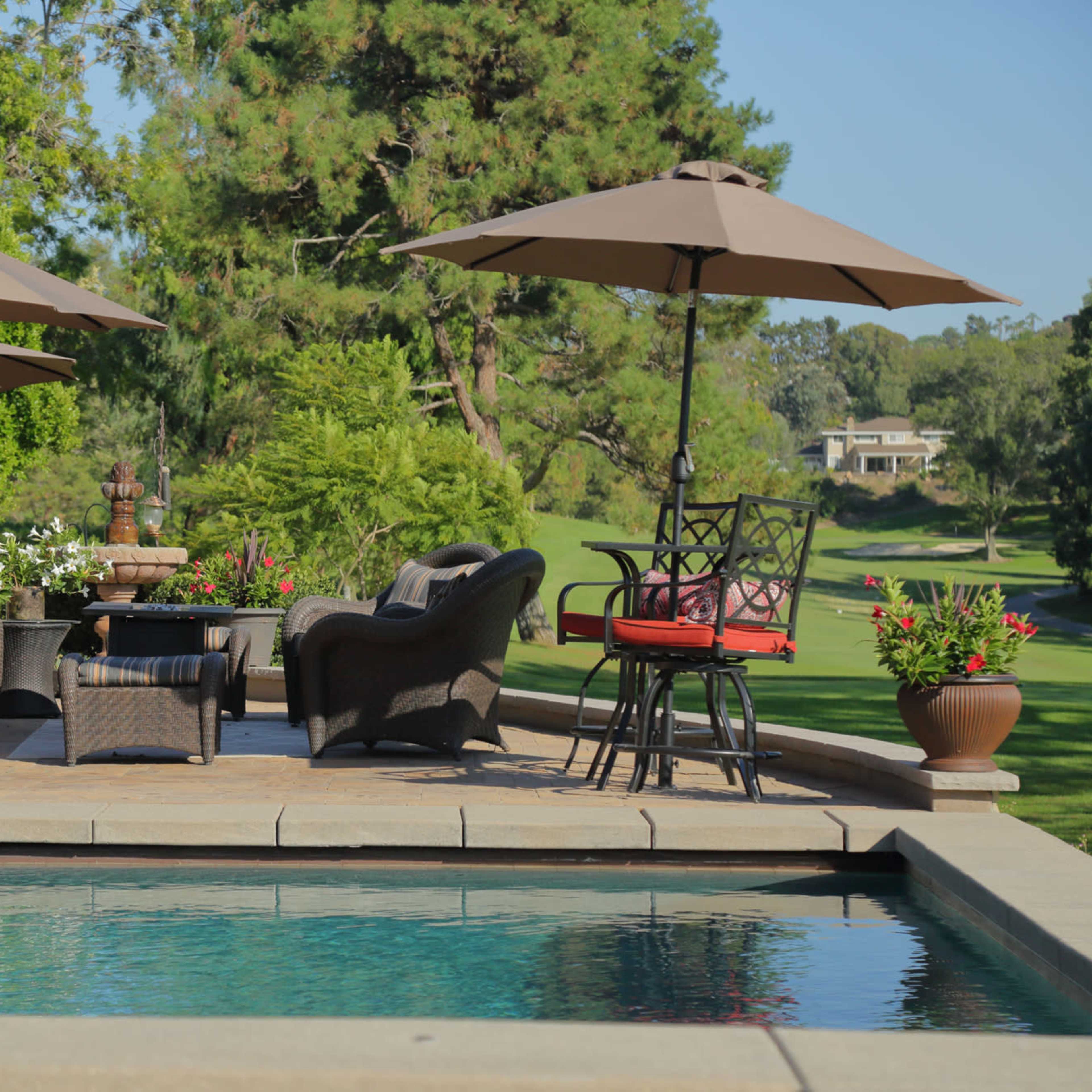 A patio area features wicker furniture and umbrellas alongside a swimming pool, surrounded by landscaped greenery.