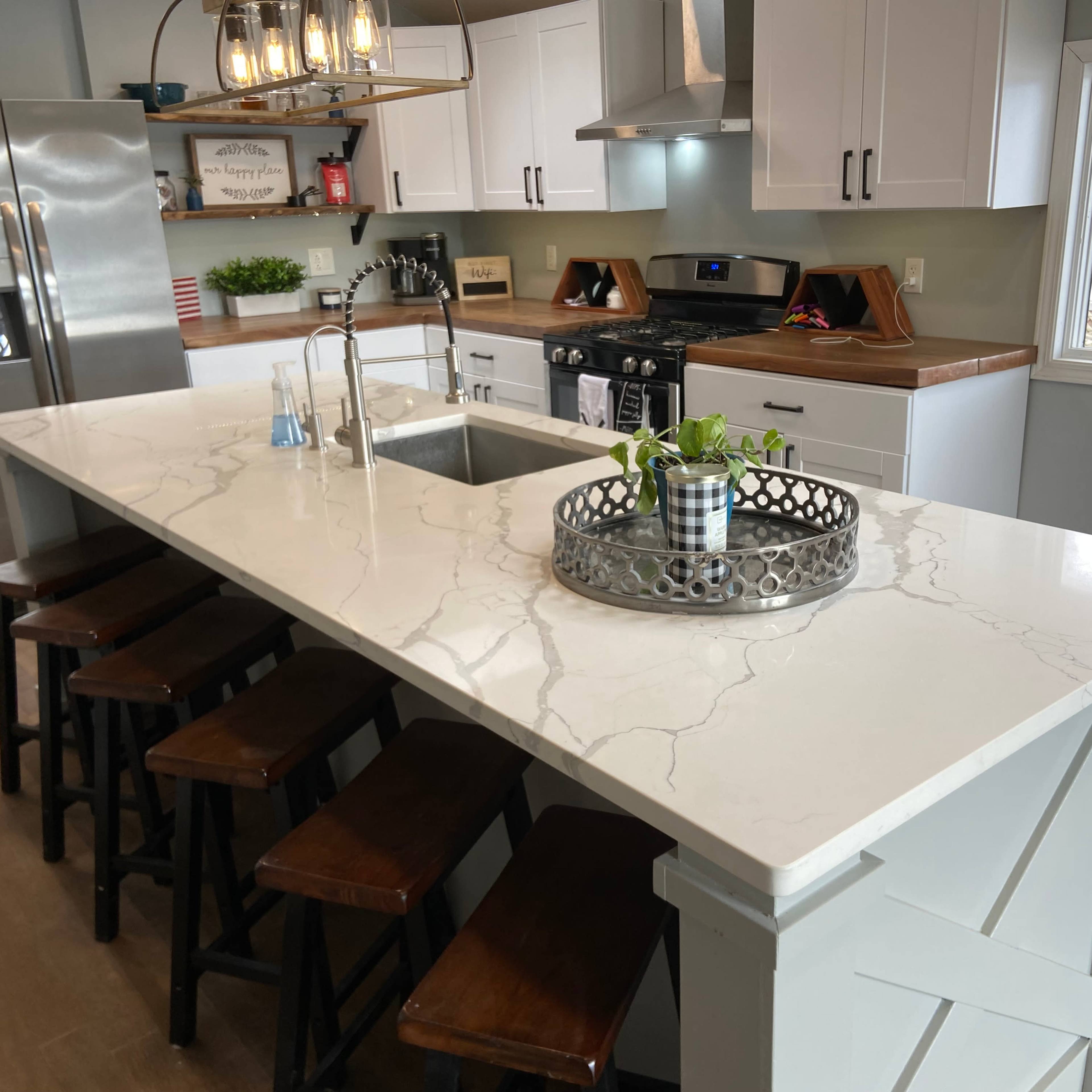 A modern kitchen features a large white countertop with a sink, surrounded by dark wooden stools, and equipped with stainless steel appliances.