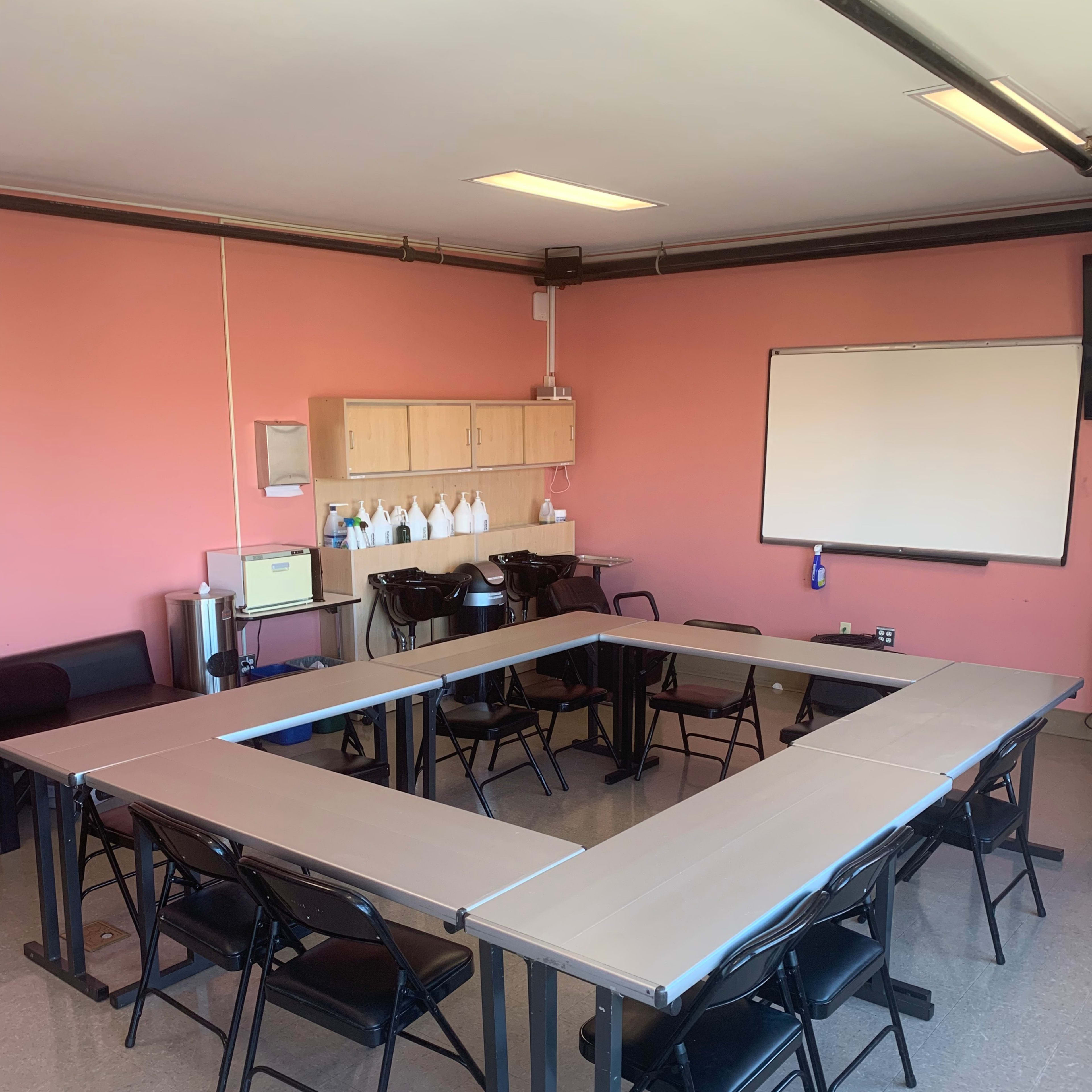 A meeting room features a rectangular arrangement of tables surrounded by black chairs, with a pink wall and a whiteboard in the background.