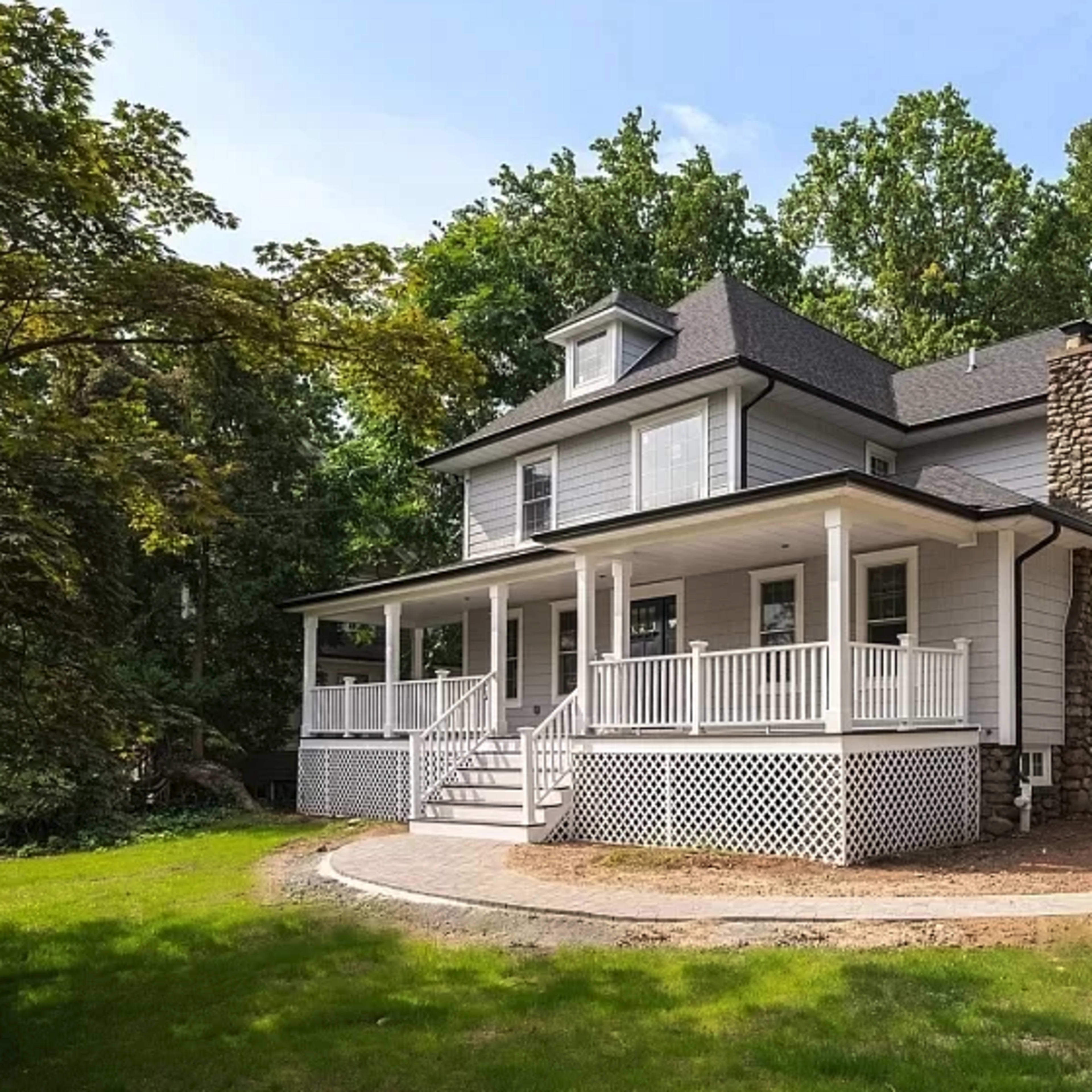 A two-story house with a wraparound porch, surrounded by greenery and a stone pathway.