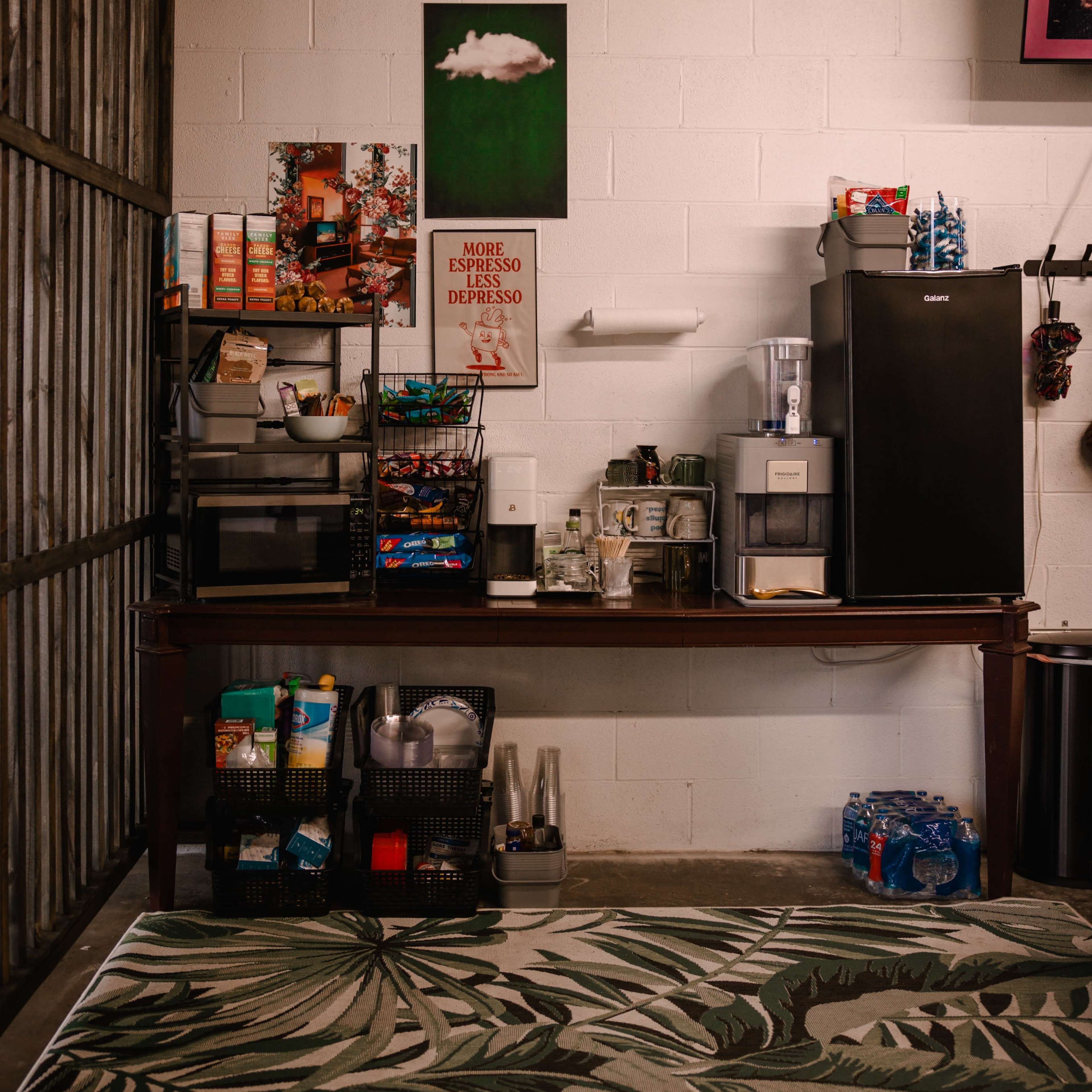 A workspace with a large table featuring various kitchen supplies, a coffee maker, and a refrigerator against a wall decorated with a green artwork and a motivational poster.