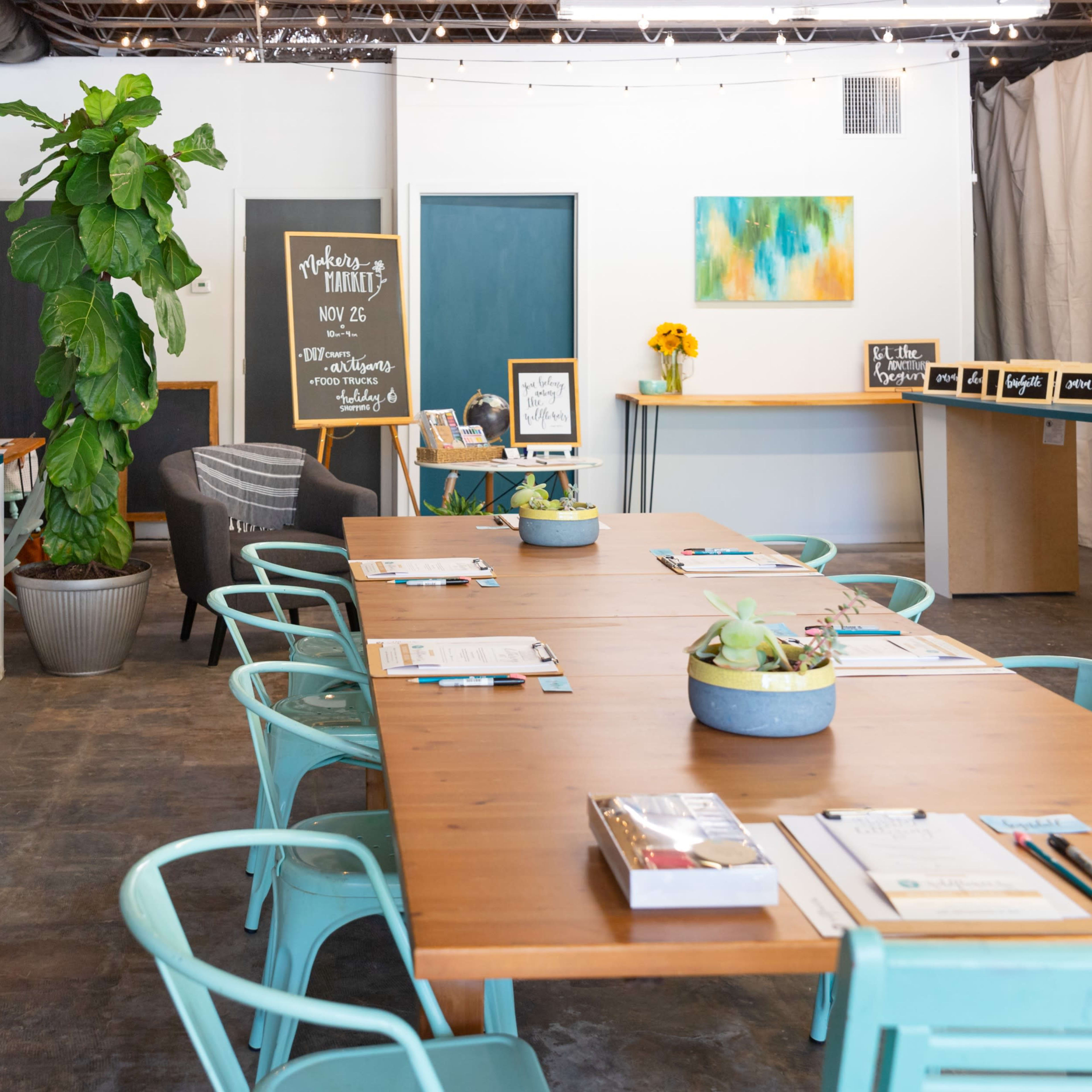 A well-lit, modern workspace features a long wooden table surrounded by teal chairs, with a variety of plants and decorative items in the background.