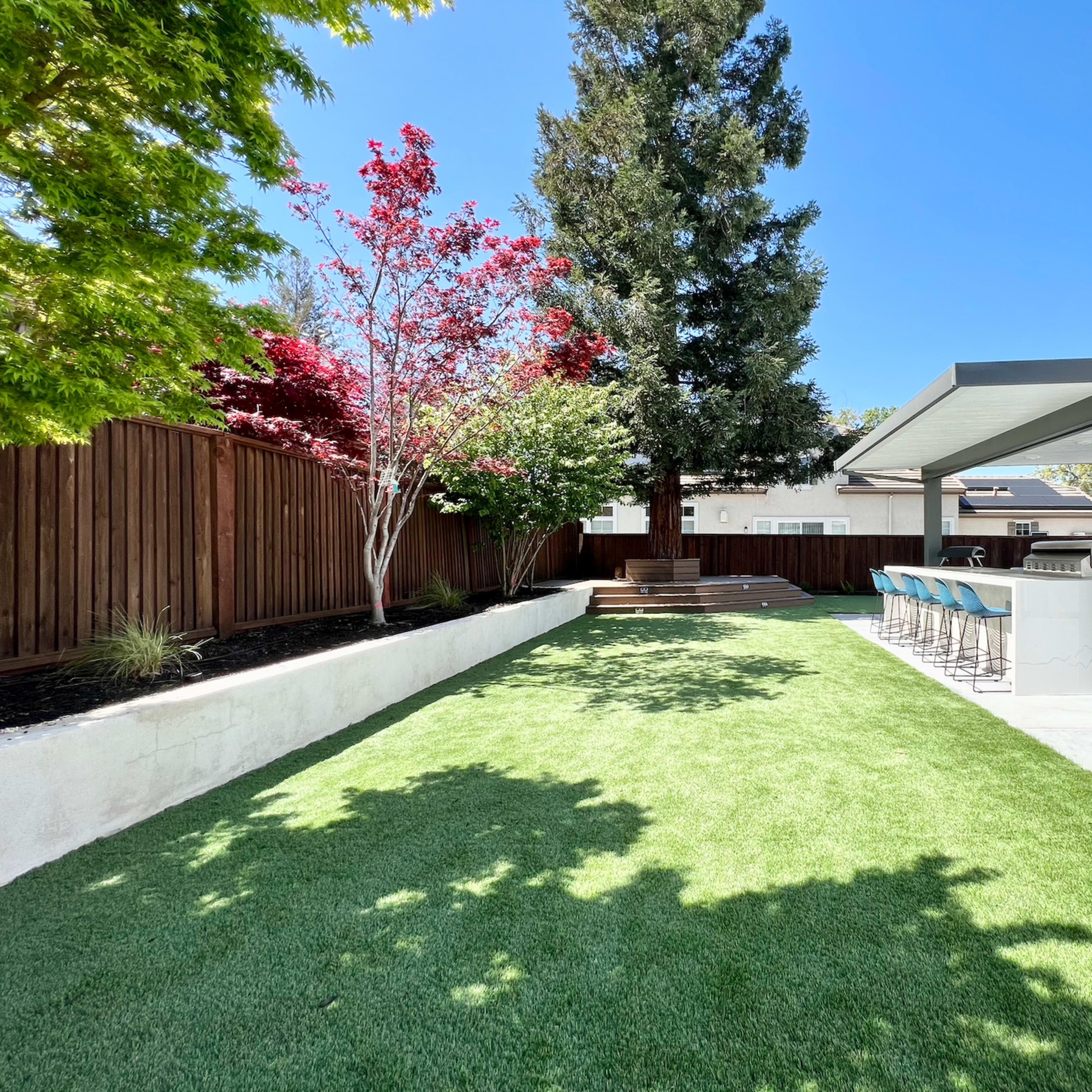 A landscaped backyard featuring artificial grass, a dining area with bar stools, and several trees along a wooden fence.
