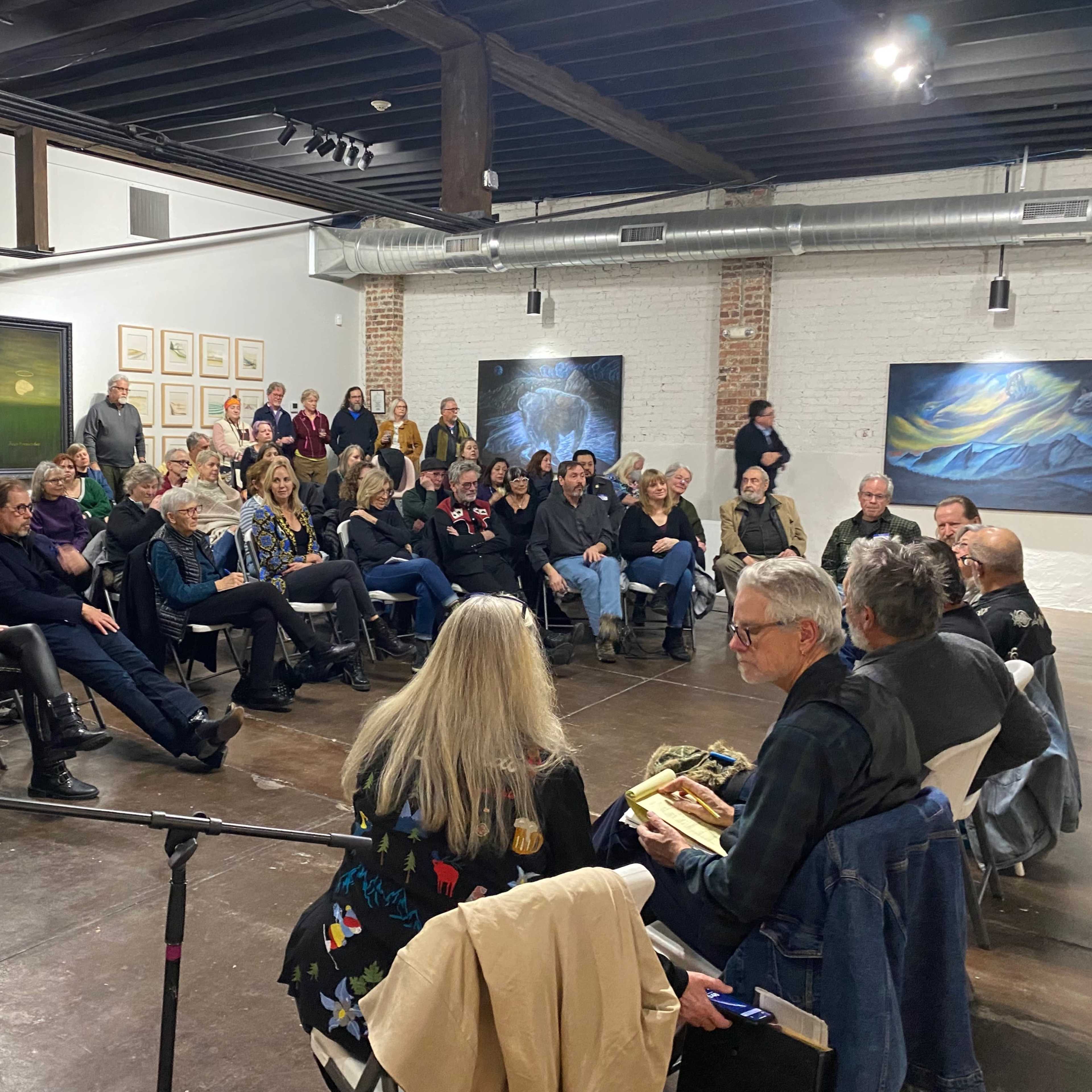 A crowd of people is seated in a gallery space, attentively listening to speakers at a discussion event.