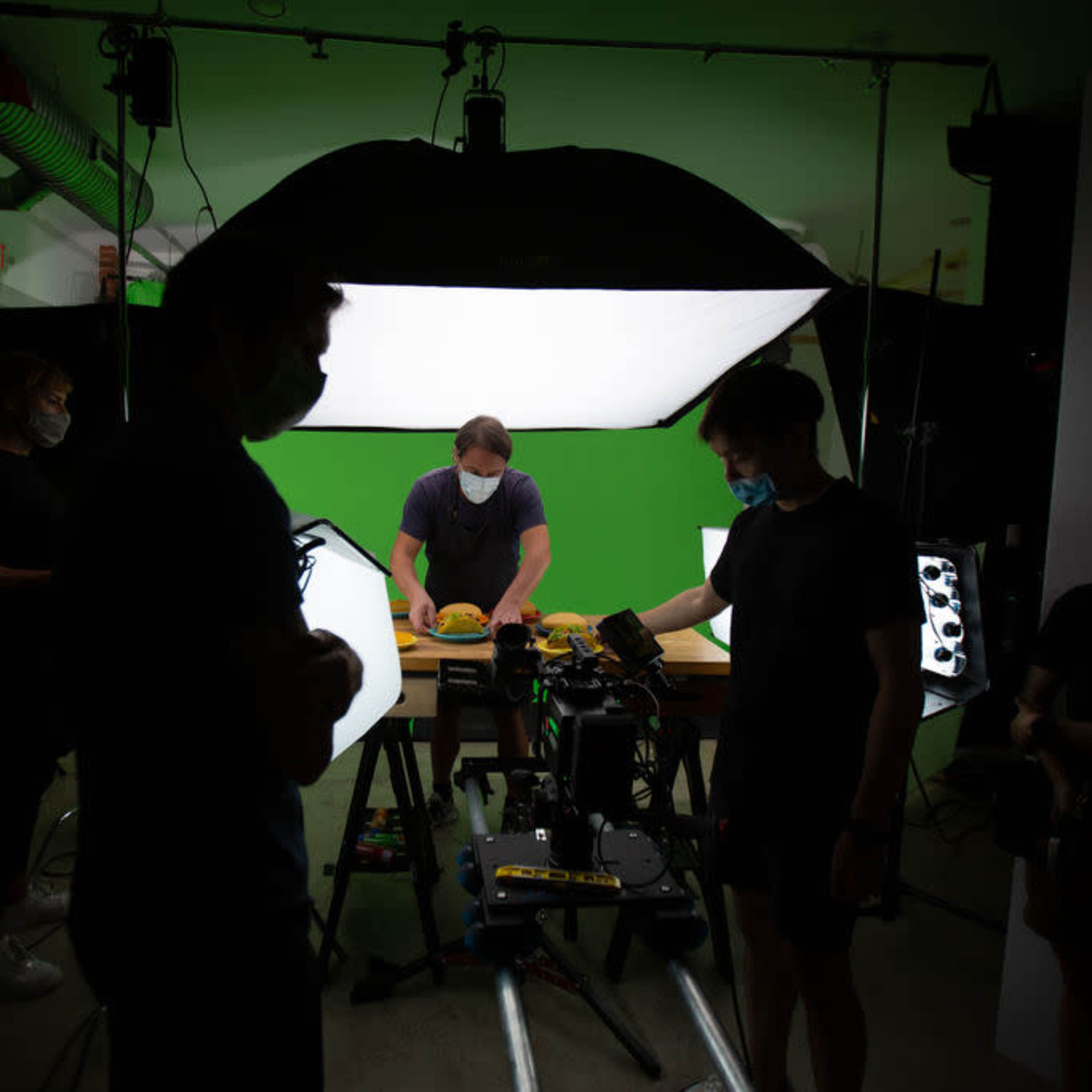 A film crew sets up lighting and equipment in a studio with a green screen backdrop.