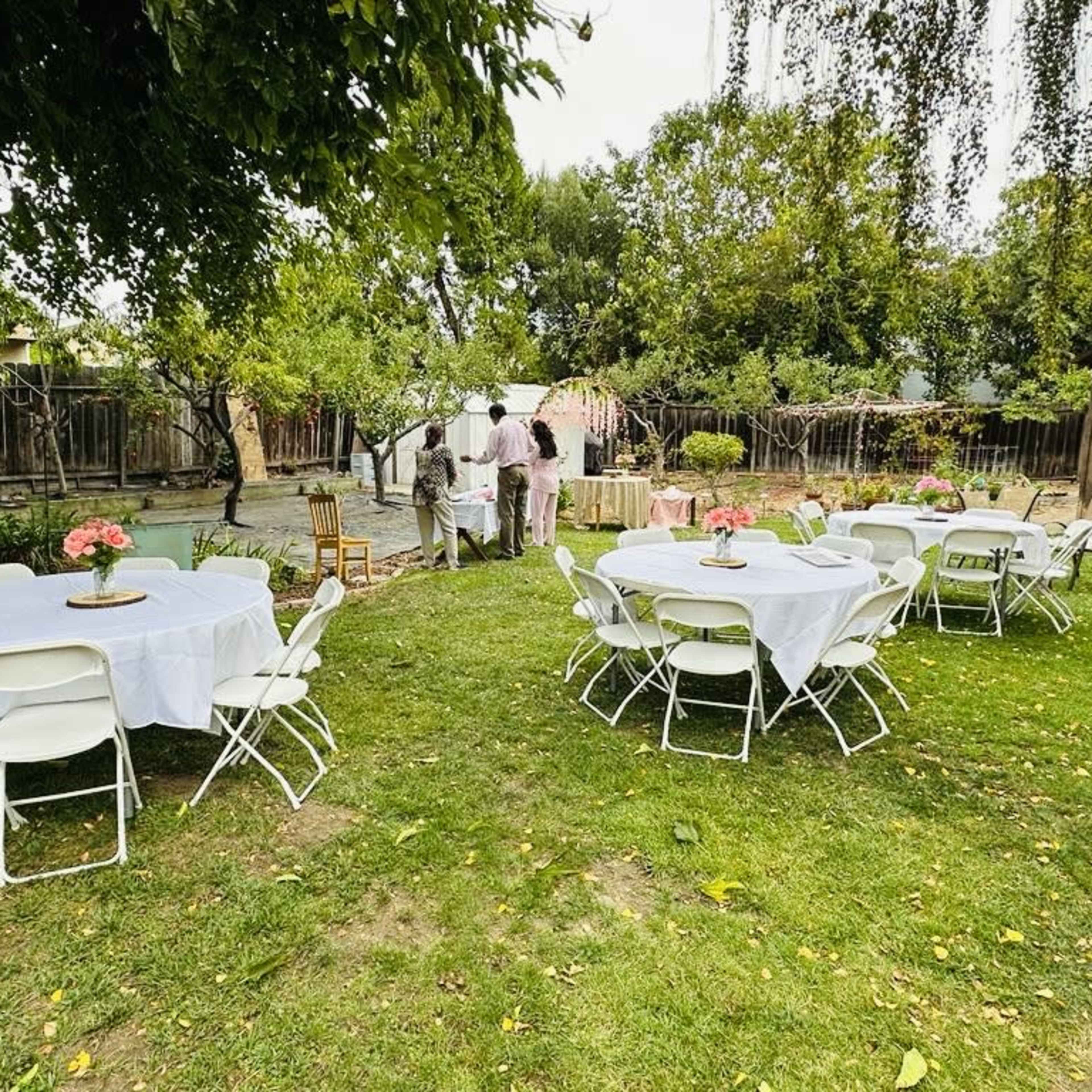 A backyard event setup with round tables covered in white tablecloths and floral centerpieces, surrounded by greenery and a wooden fence.