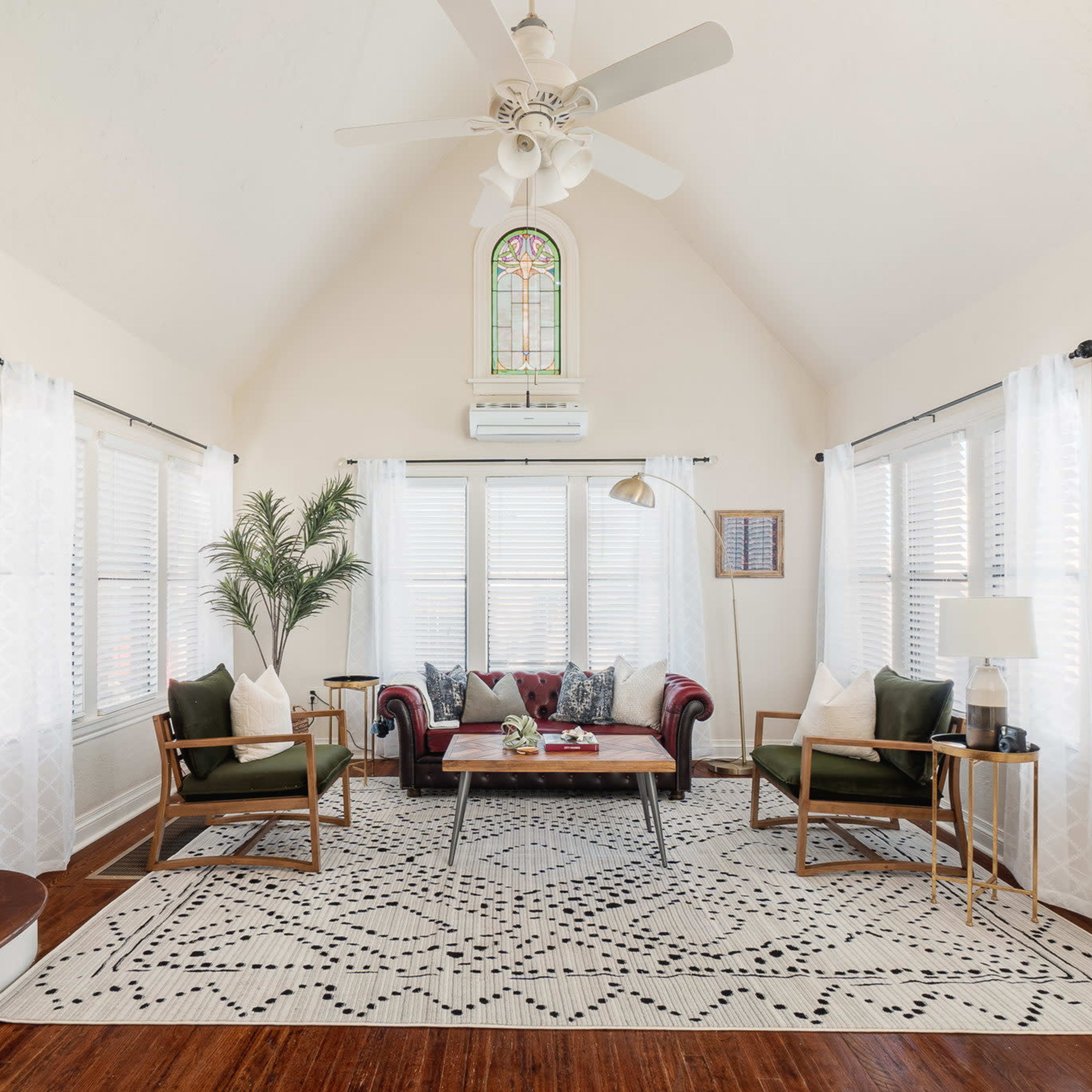 A bright living room with a high ceiling, featuring a red couch, green armchairs, a coffee table, and large windows adorned with light curtains.
